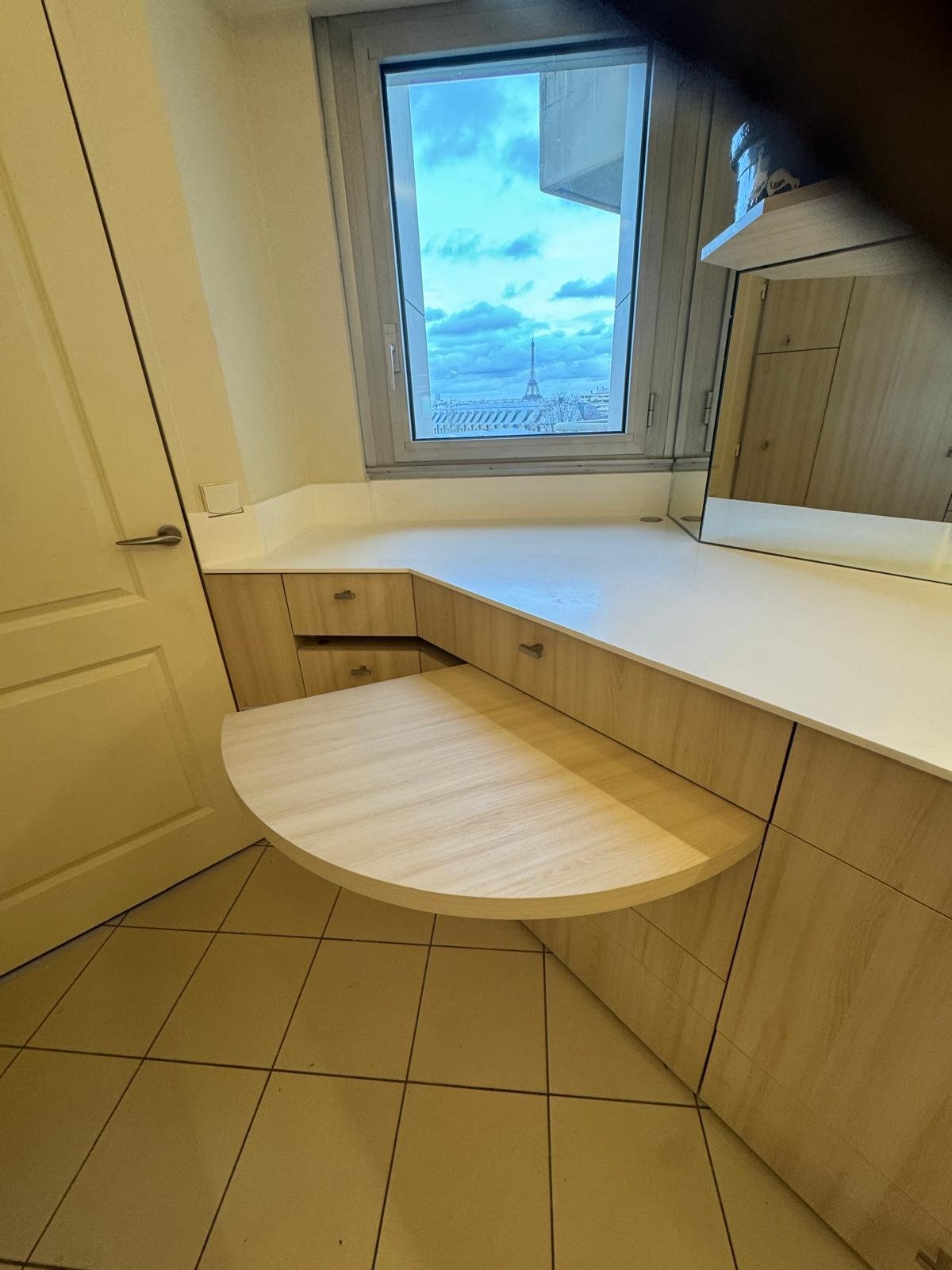 A corner of a room with a window showing a cloudy sky and the Eiffel Tower in the distance, a built-in wooden desk with drawers, and a mirror on the wall.