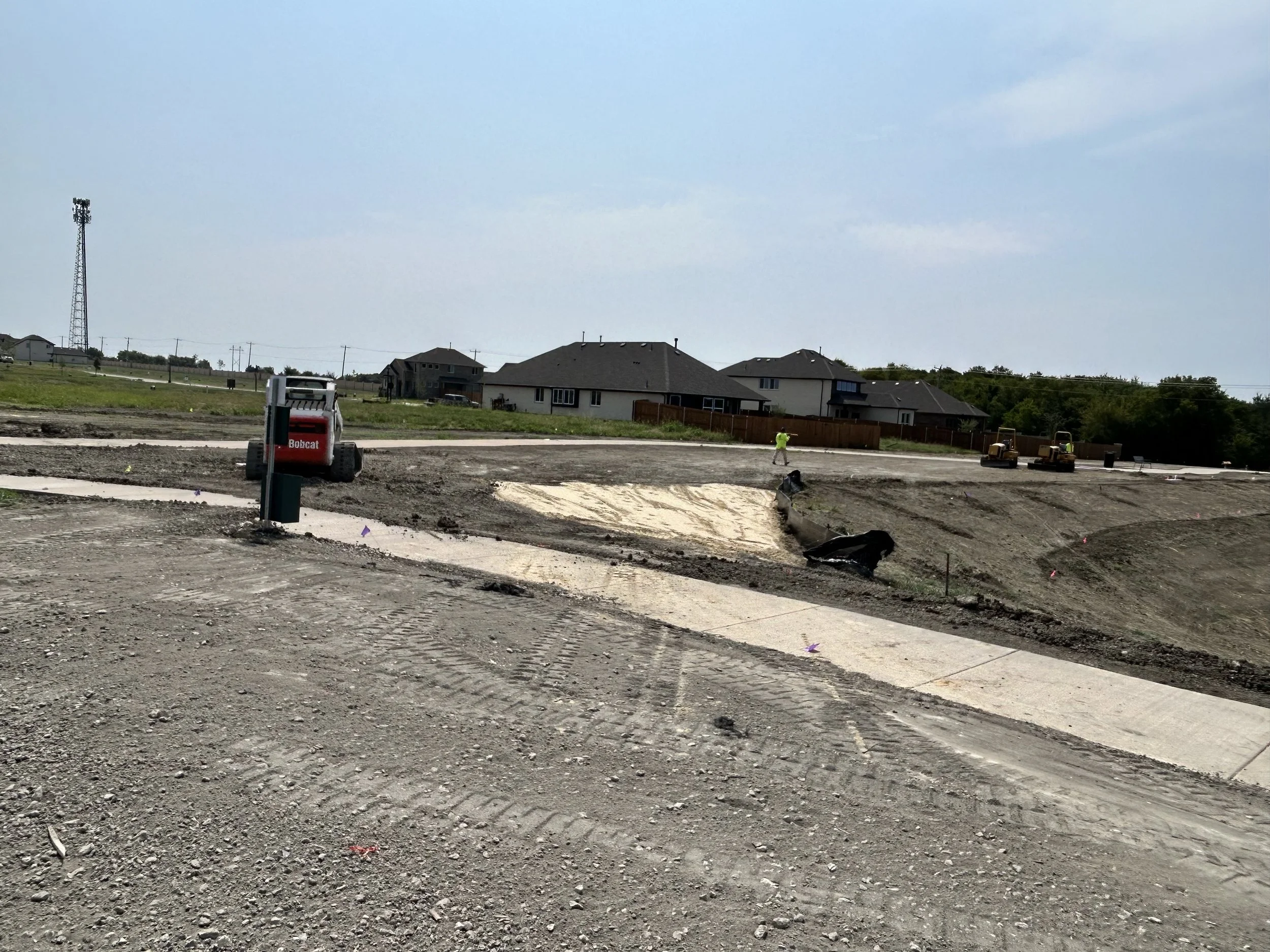Construction site with uneven ground, wheelbarrow, and workers. Houses in background under a cloudy sky.