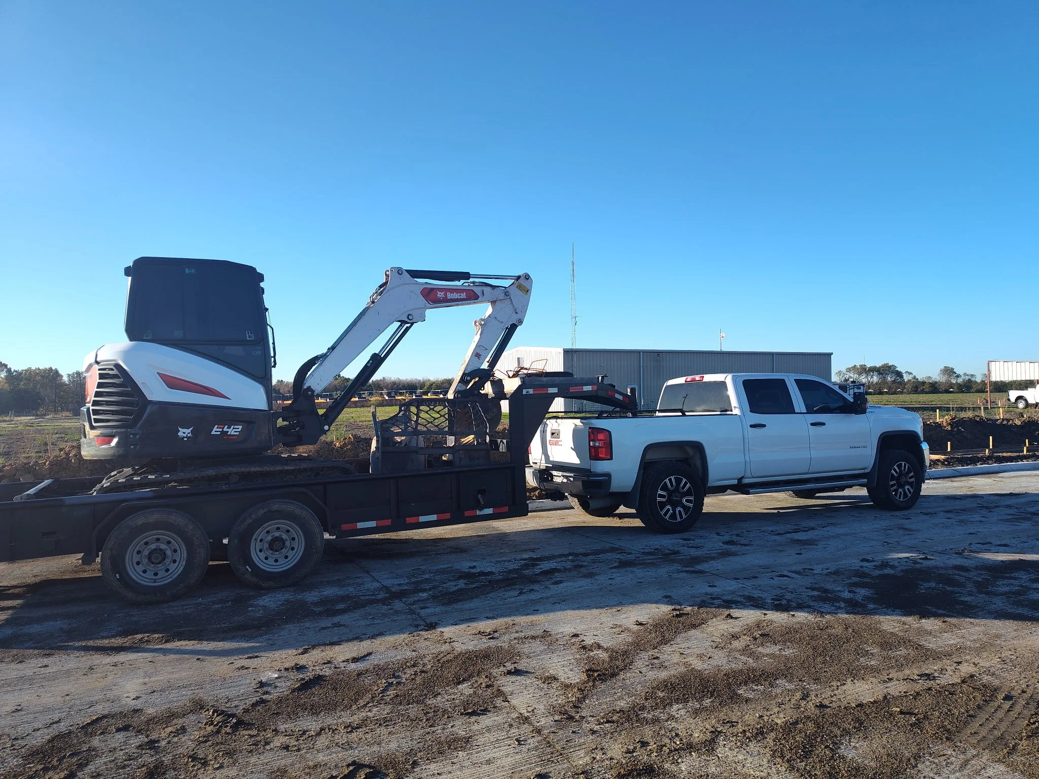A white pickup truck transporting a small excavator on a trailer at a construction site with clear blue skies and a field in the background.