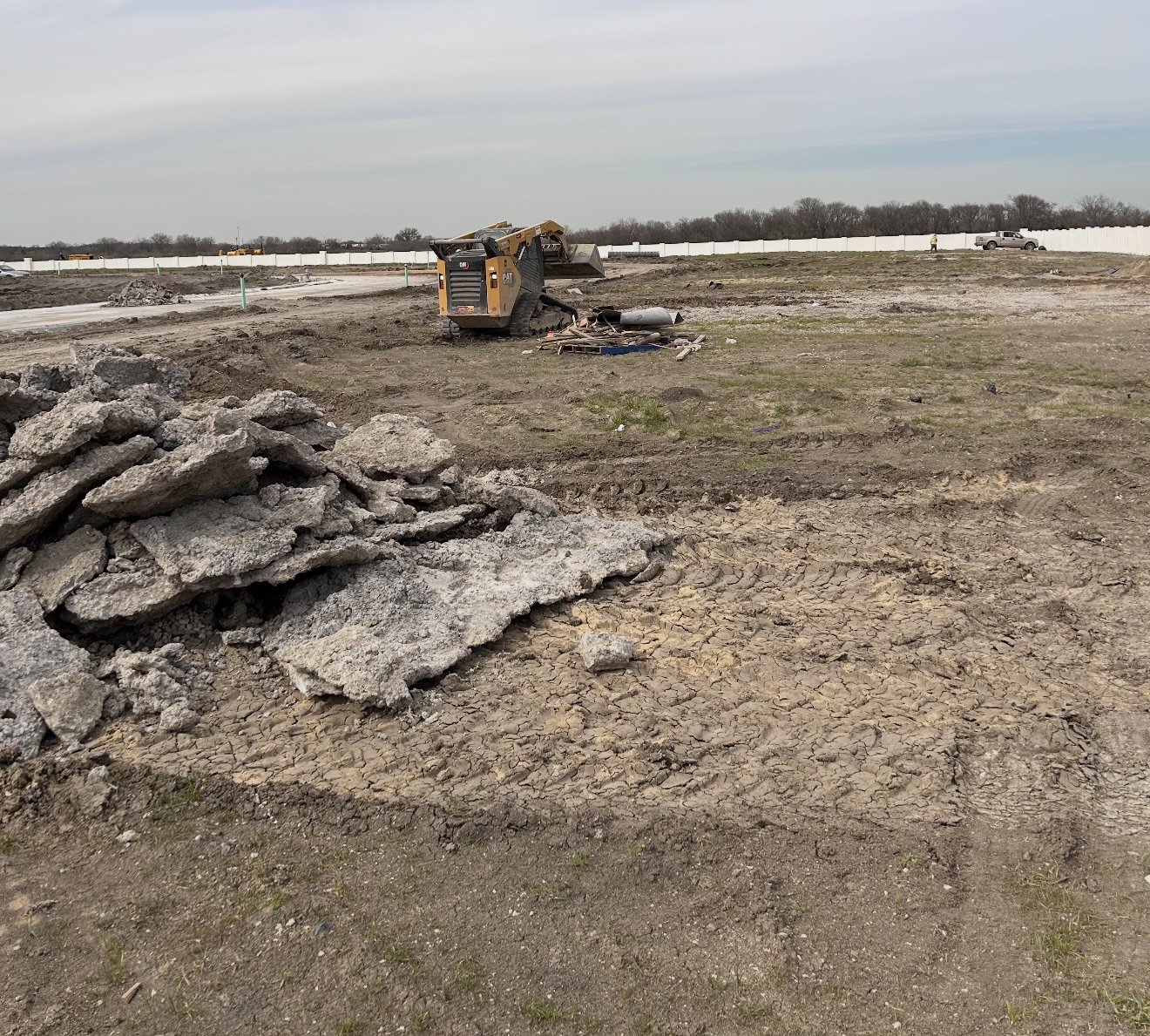 Construction site with a small bulldozer and a pile of broken concrete debris, patches of dirt, and some construction tools scattered on the ground. Fencing and vehicles are visible in the background.