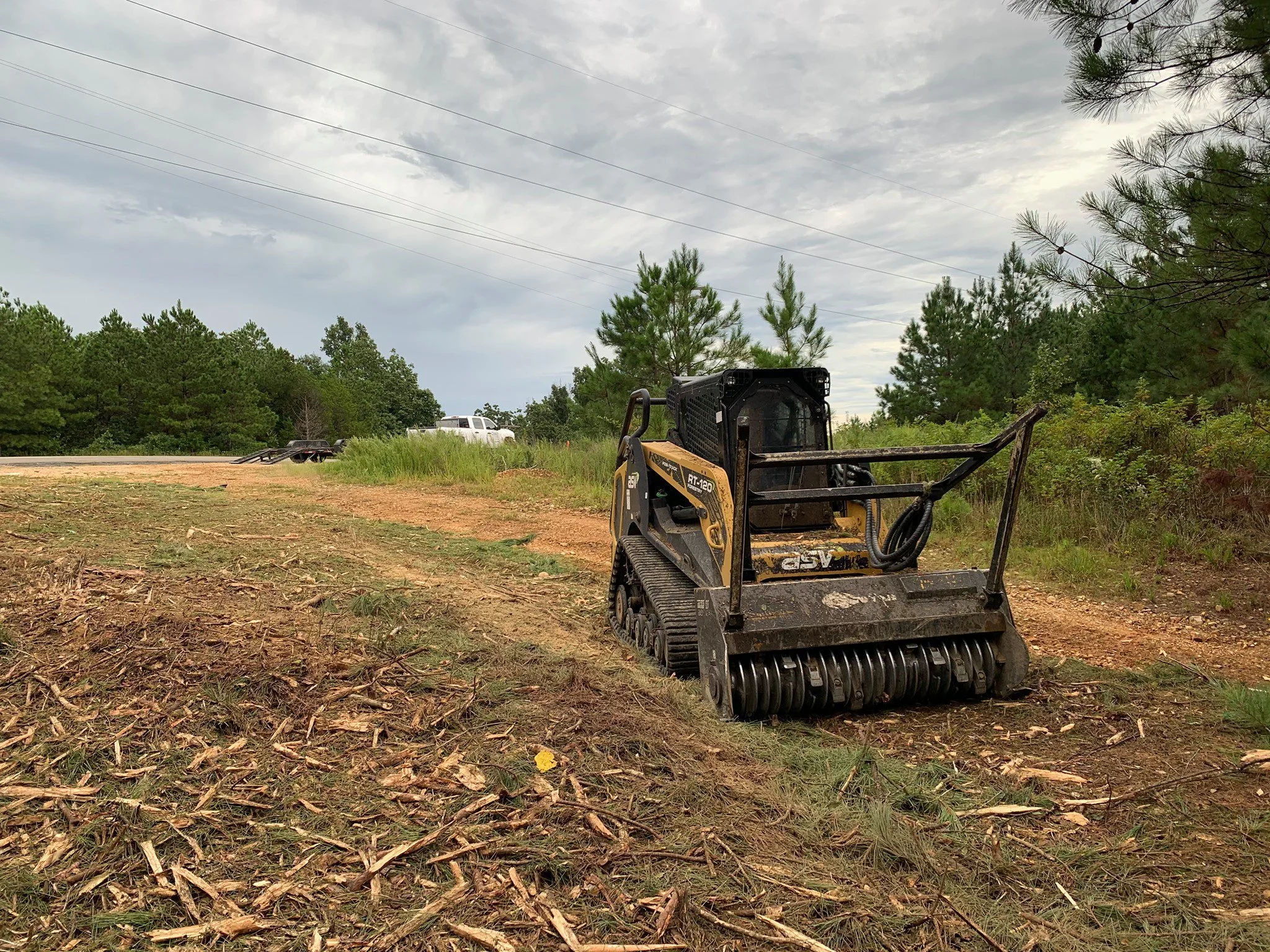 A small yellow and black skid steer loader on a dirt patch with tire tracks, surrounded by green trees and under a cloudy sky.