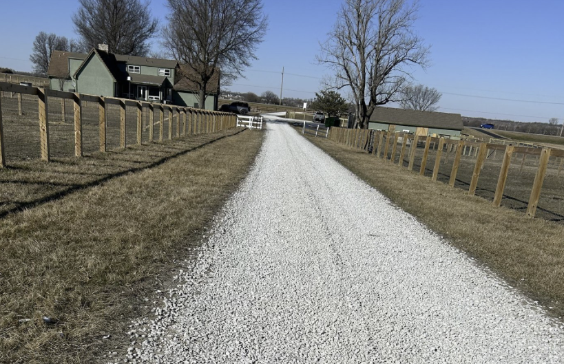 A gravel driveway flanked by grassy areas and wooden fences, leading to houses in the distance on a clear, sunny day.