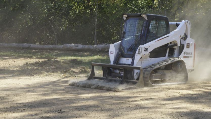 A Bobcat compact track loader clearing dirt and dust on a construction site with trees in the background.