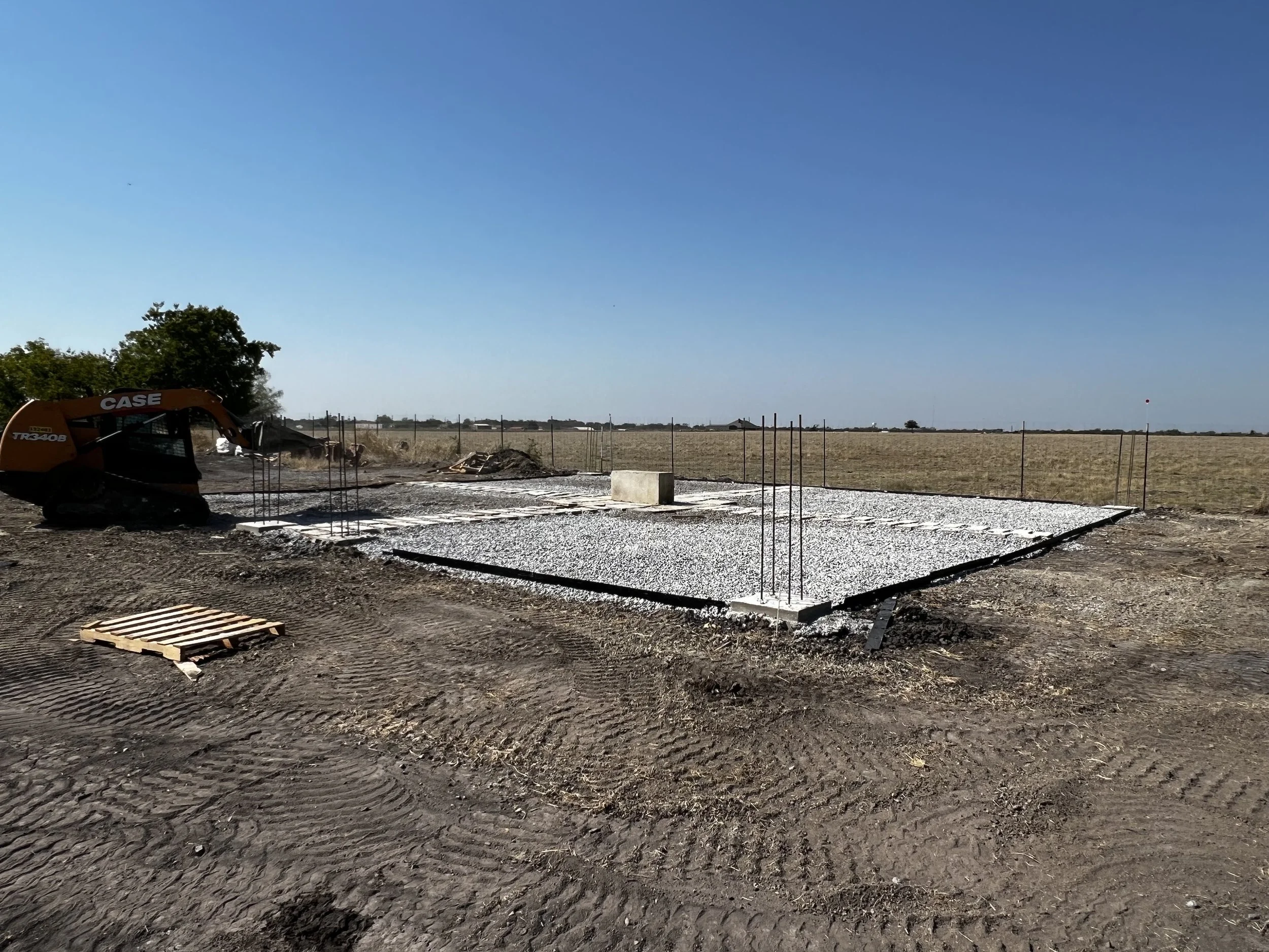 Construction site with a small excavator, gravel foundation, and rebar sticking out for a building to be built on a rural property under a clear blue sky.