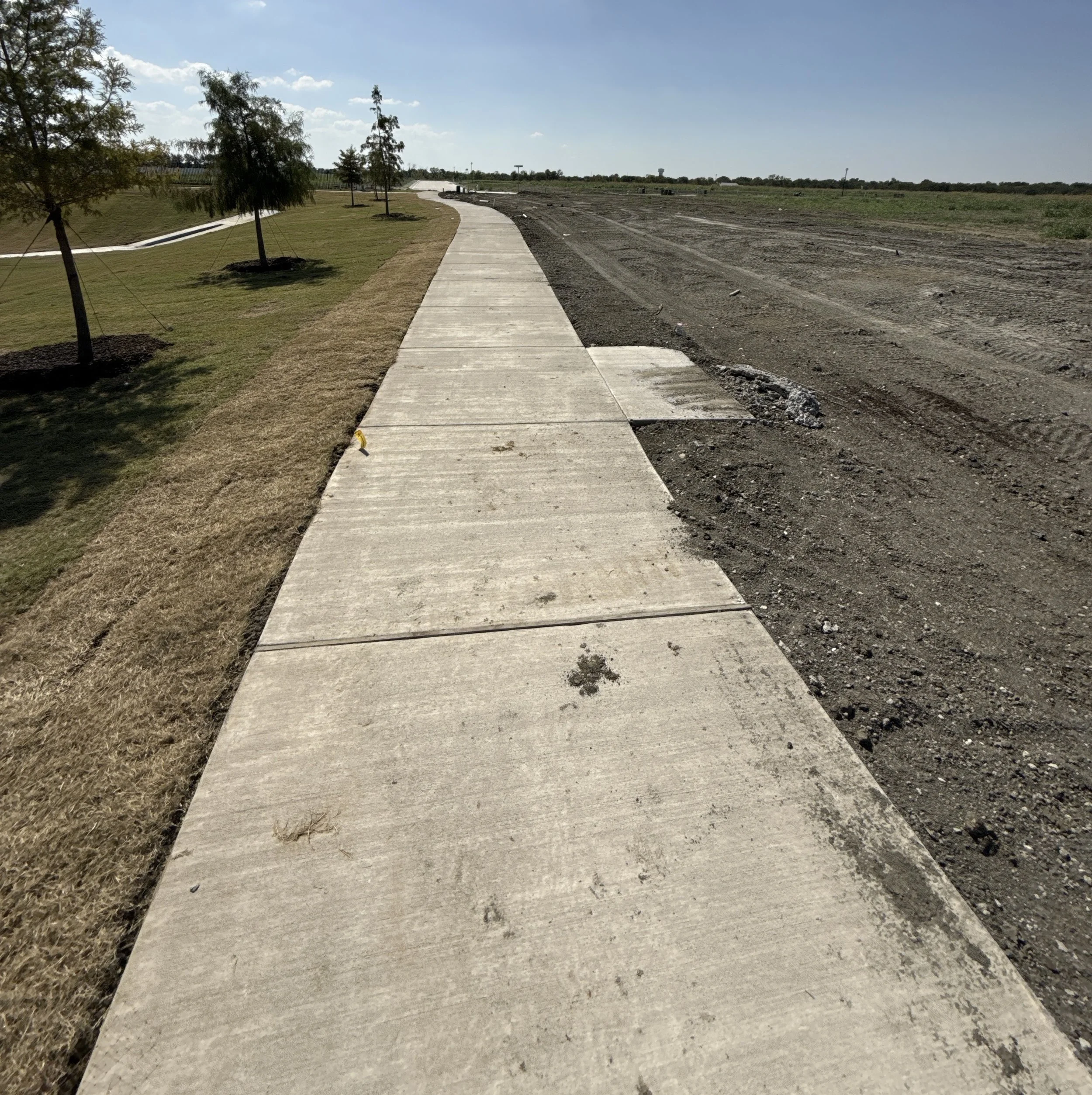 Newly constructed concrete sidewalk alongside an area of grass and trees on the left, with dirt and construction work on the right, under a partly cloudy sky.
