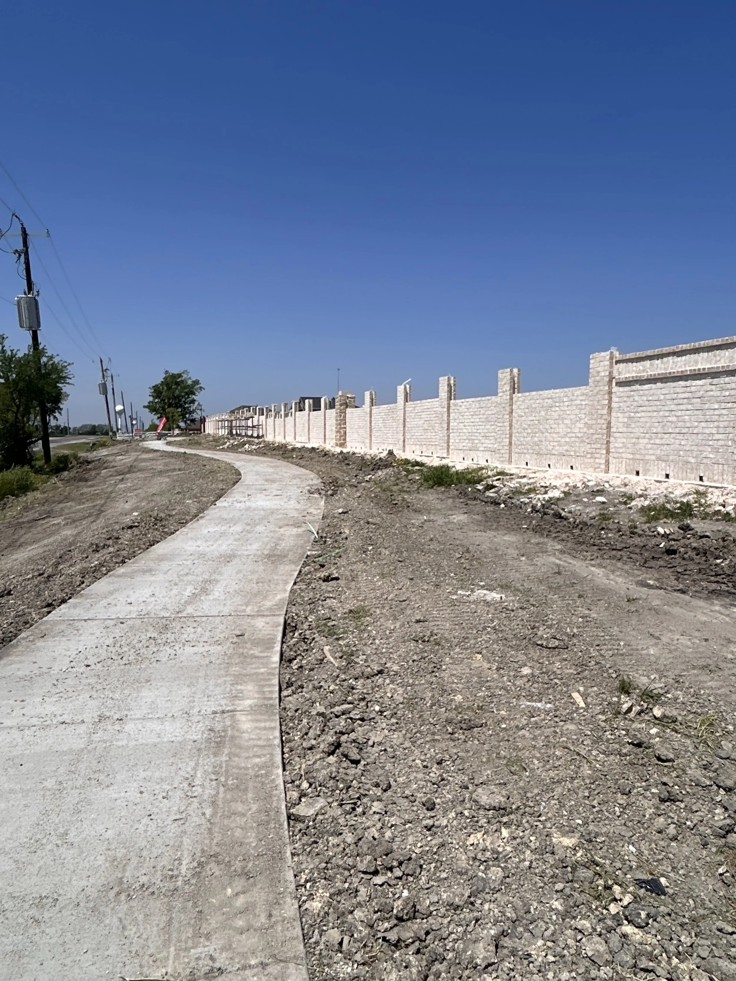 A winding sidewalk alongside a brick wall under a clear blue sky, with utility poles and some trees visible in the distance.