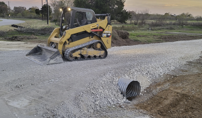 Small construction vehicle (compact track loader) parked on a gravel road, with a large corrugated pipe partially buried in the ground nearby, under a sunset-lit sky.