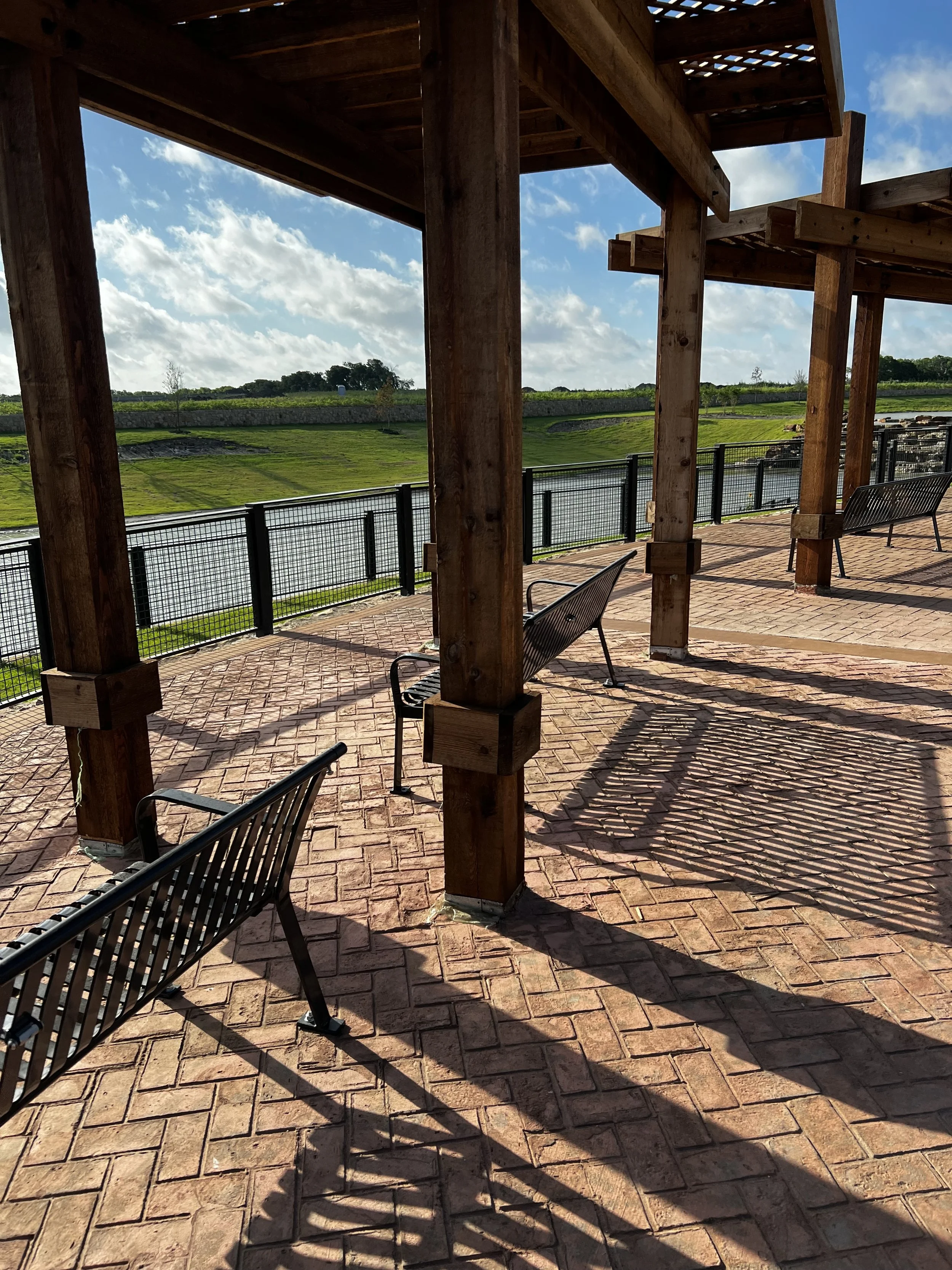 Empty wooden shelter with metal benches on brick-paved ground overlooking a water and green landscape with cloudy sky.