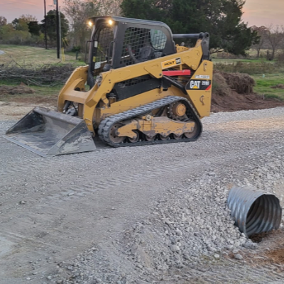 A yellow compact track loader with a black safety cage operates on a gravel surface at a construction site, with a metal drainage pipe on the ground nearby.