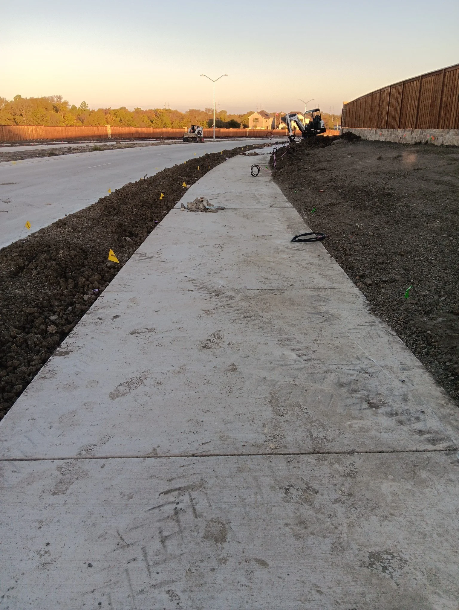 Newly constructed sidewalk on a road, with construction equipment and materials visible in the background, and a fence on the right side.