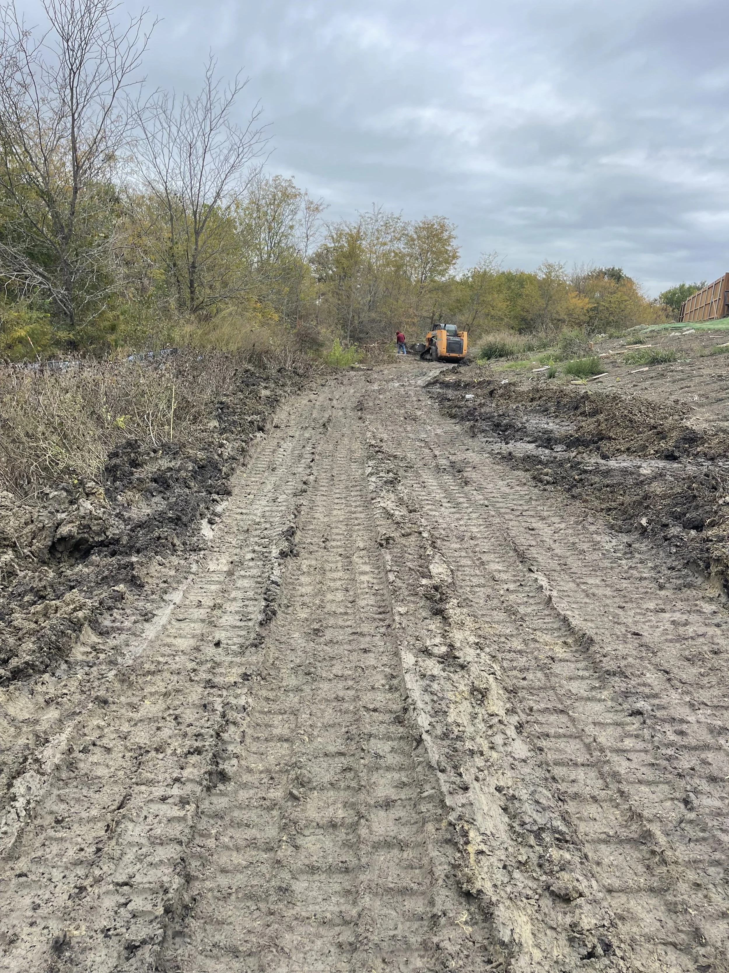 A dirt construction road with tire tracks, leading to an orange compactor machine and two workers in the distance under a cloudy sky, with trees on both sides.
