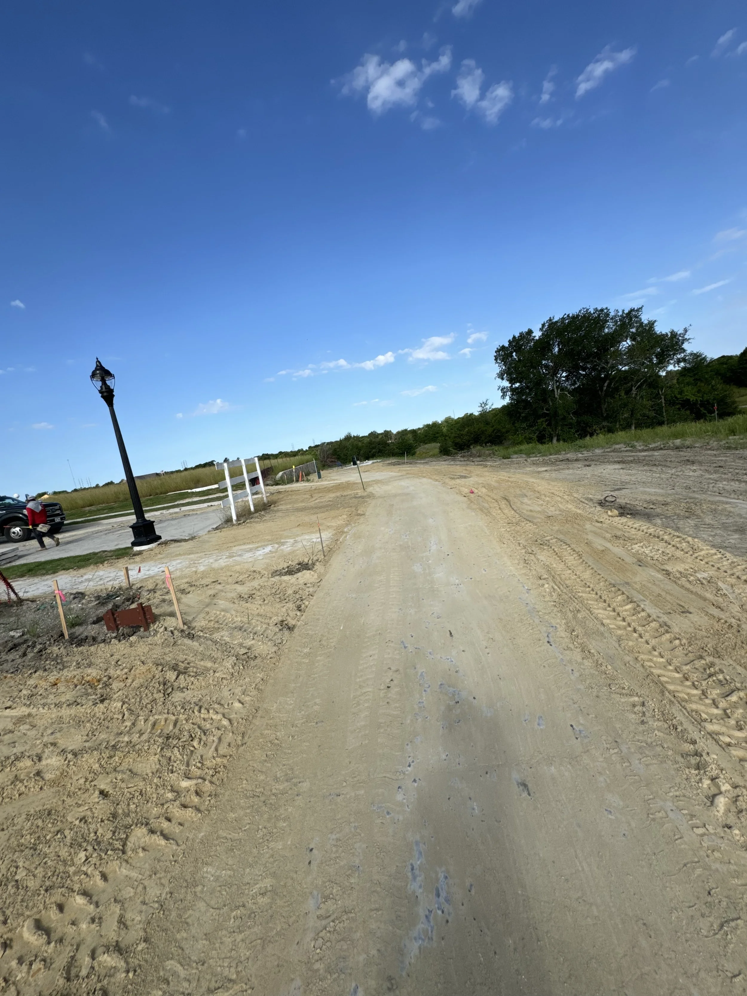 A dirt pathway under a blue sky with some scattered clouds, lined with construction markers and a vintage lamppost, with a grassy area and trees in the background.