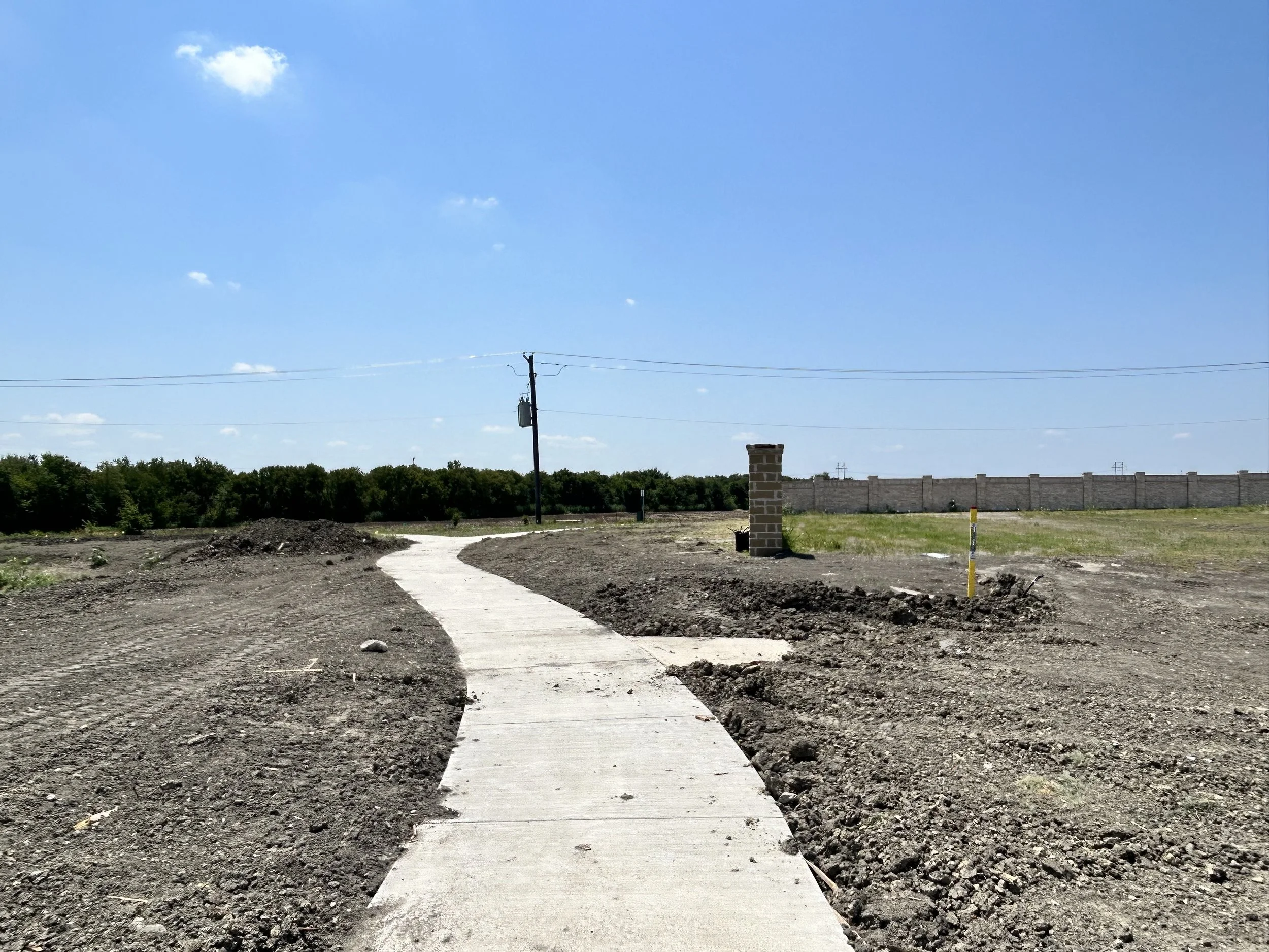 A partially constructed sidewalk winding through a barren plot of land under a bright blue sky with scattered clouds, with a utility pole and wall visible in the background.