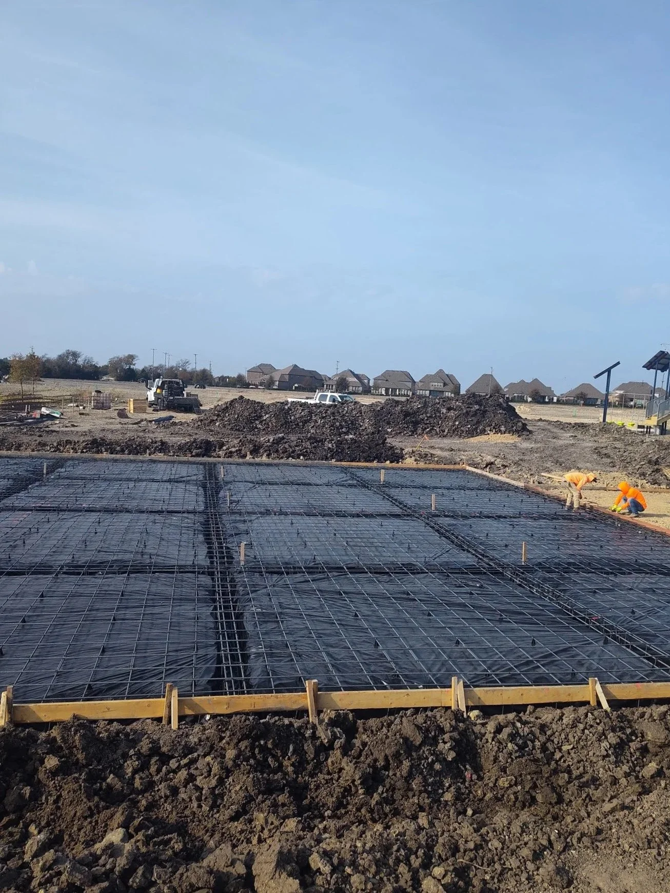 Construction site with rebar and foundation being prepared for a building, workers in orange safety vests and helmets, dirt piles, and distant houses under a blue sky. future home site, home pad, foundation prepping site prep