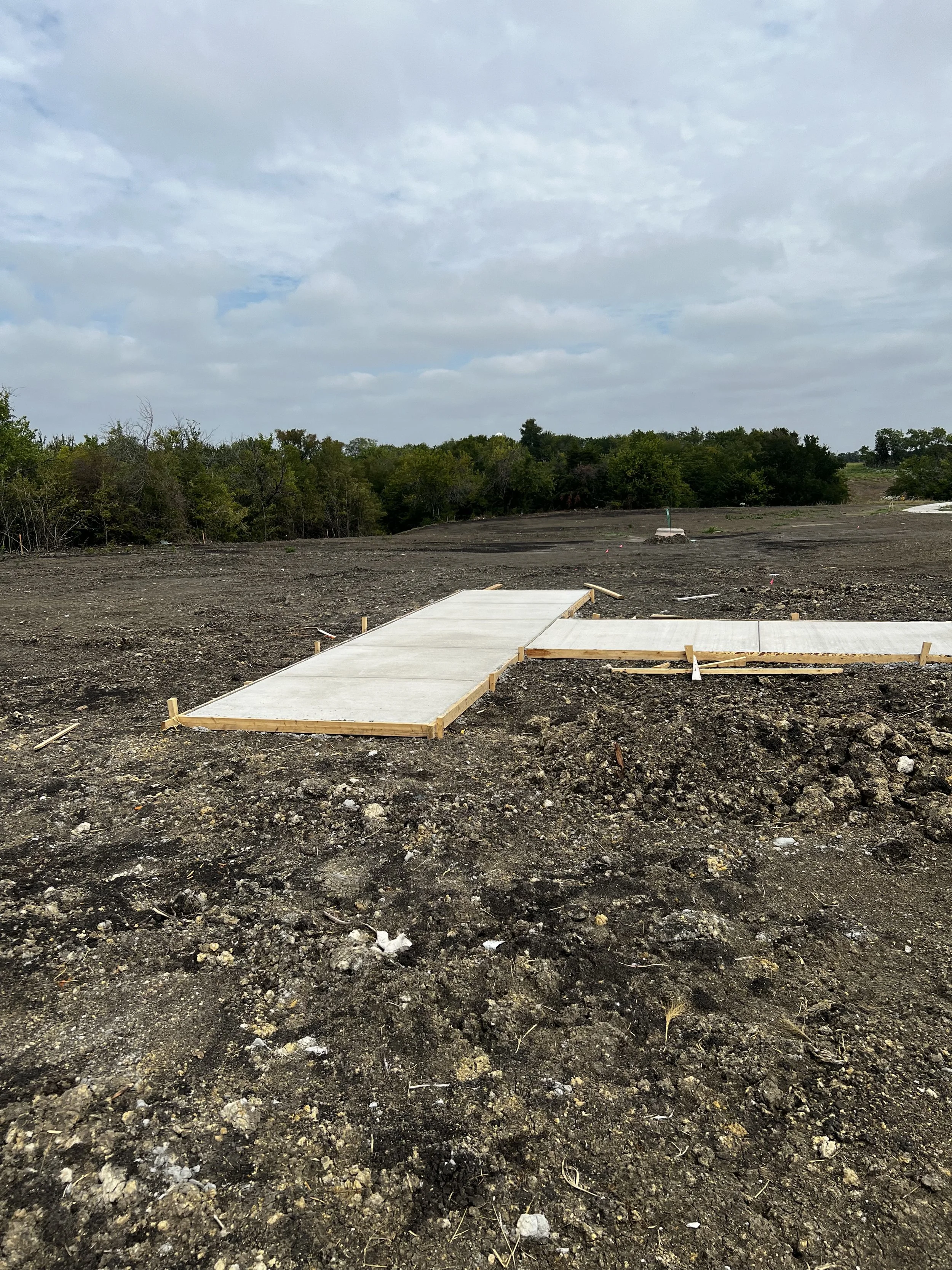 Construction site with concrete sidewalk sections being installed on dirt ground under a cloudy sky.