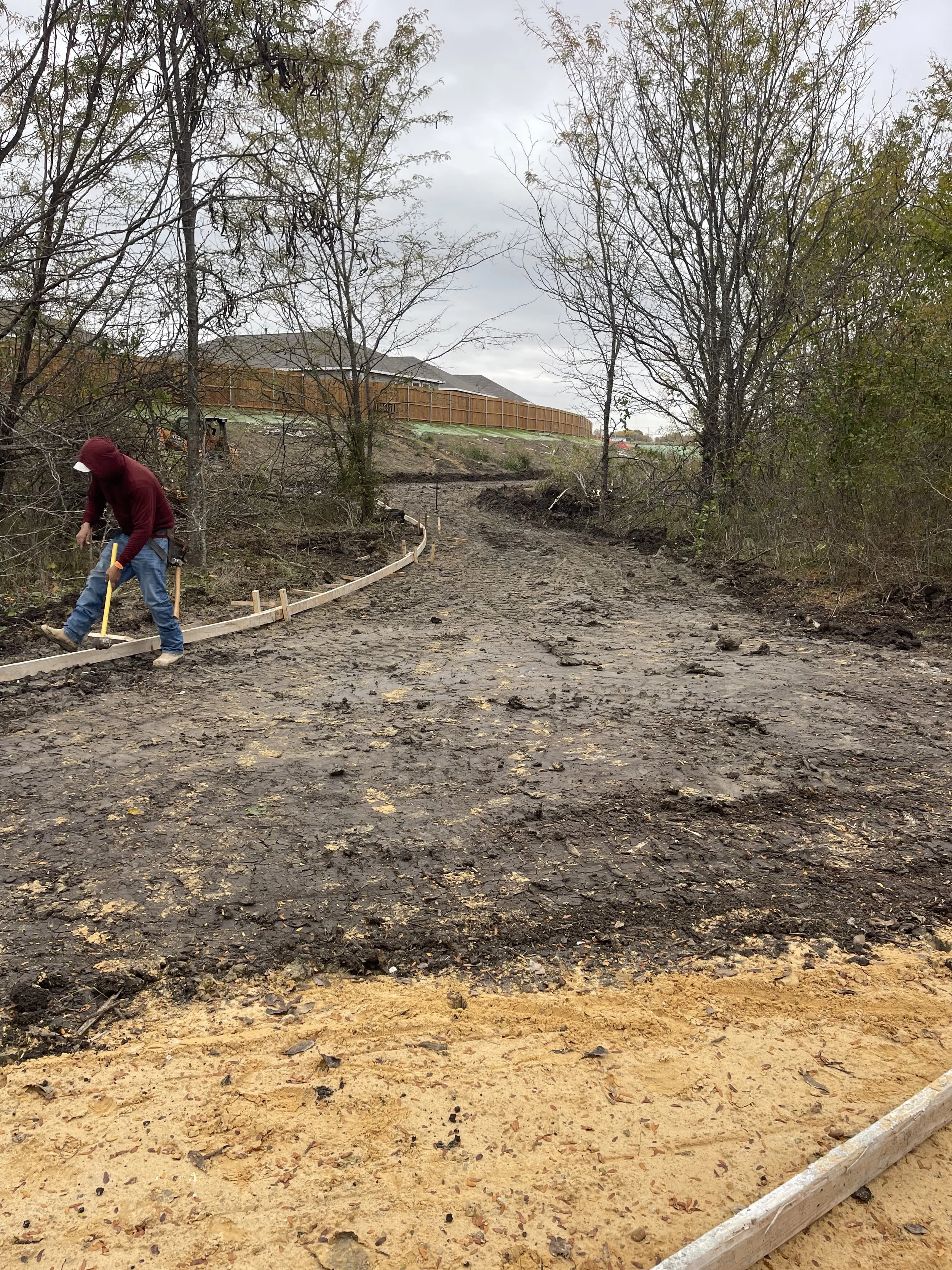 A person working on construction of a dirt path in a backyard, laying down wooden forms for the walkway, with trees and a house in the background.