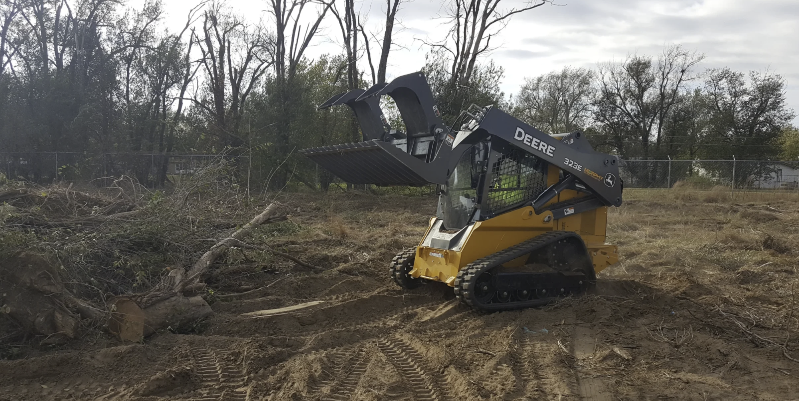 A John Deere 323E compact track loader with a grapple attachment clearing brush and debris in an outdoor construction or land clearing site.