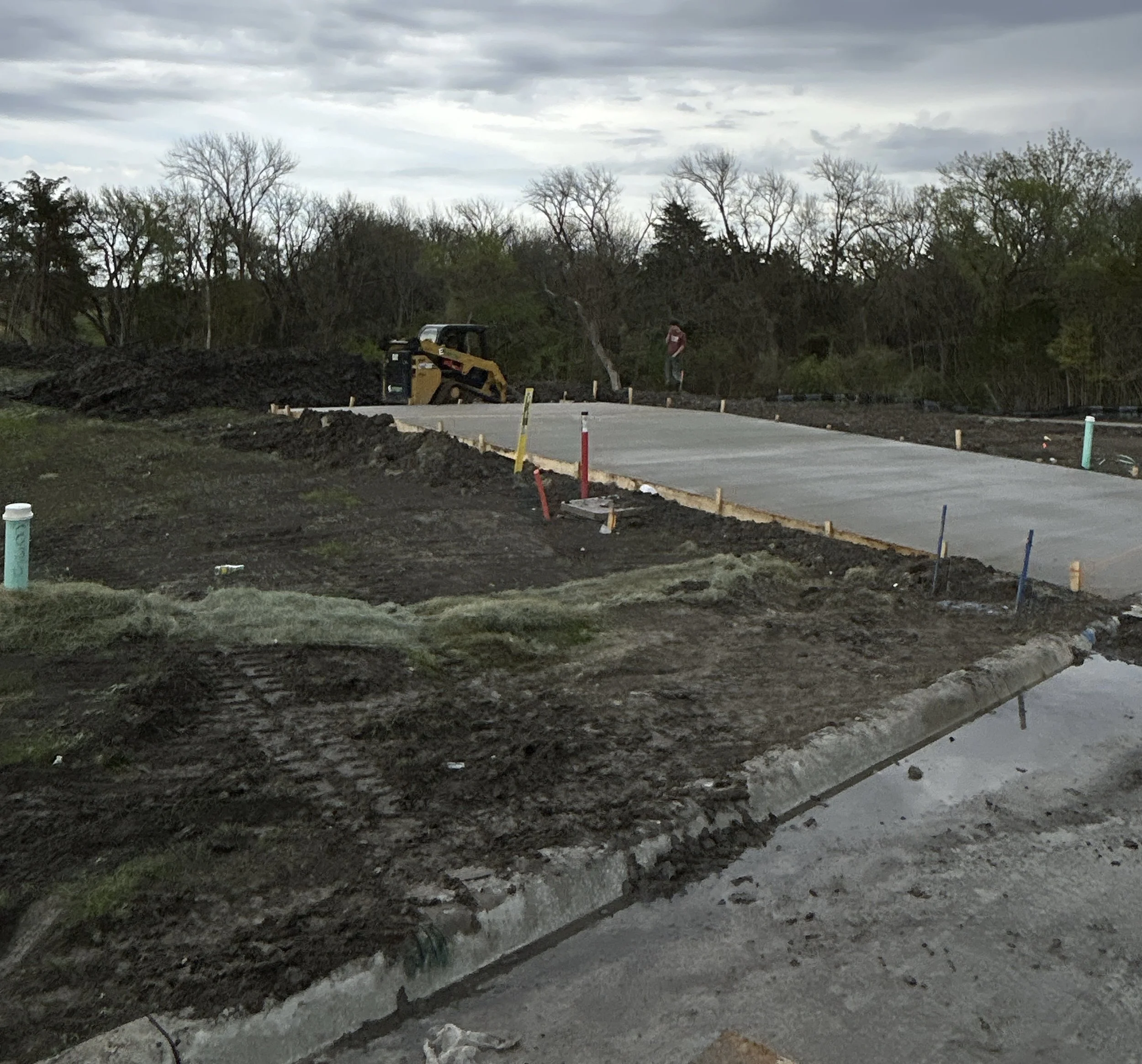 Construction site with concrete sidewalk being poured, construction equipment, and marked areas for future work, with trees and cloudy sky in the background.