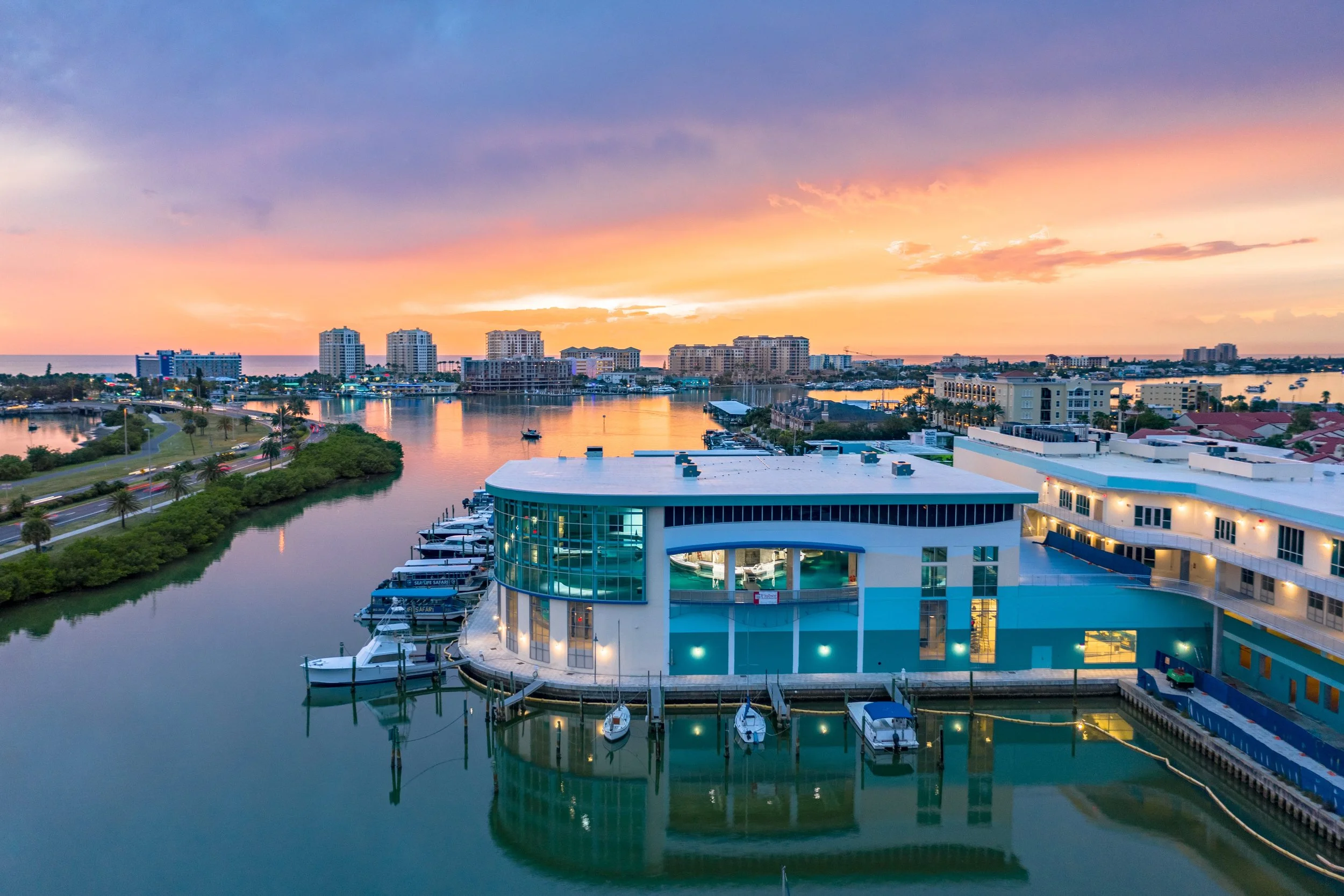 Colorful sunset over a cityscape with high-rise buildings, water, boats, and a modern waterfront building with glass windows.