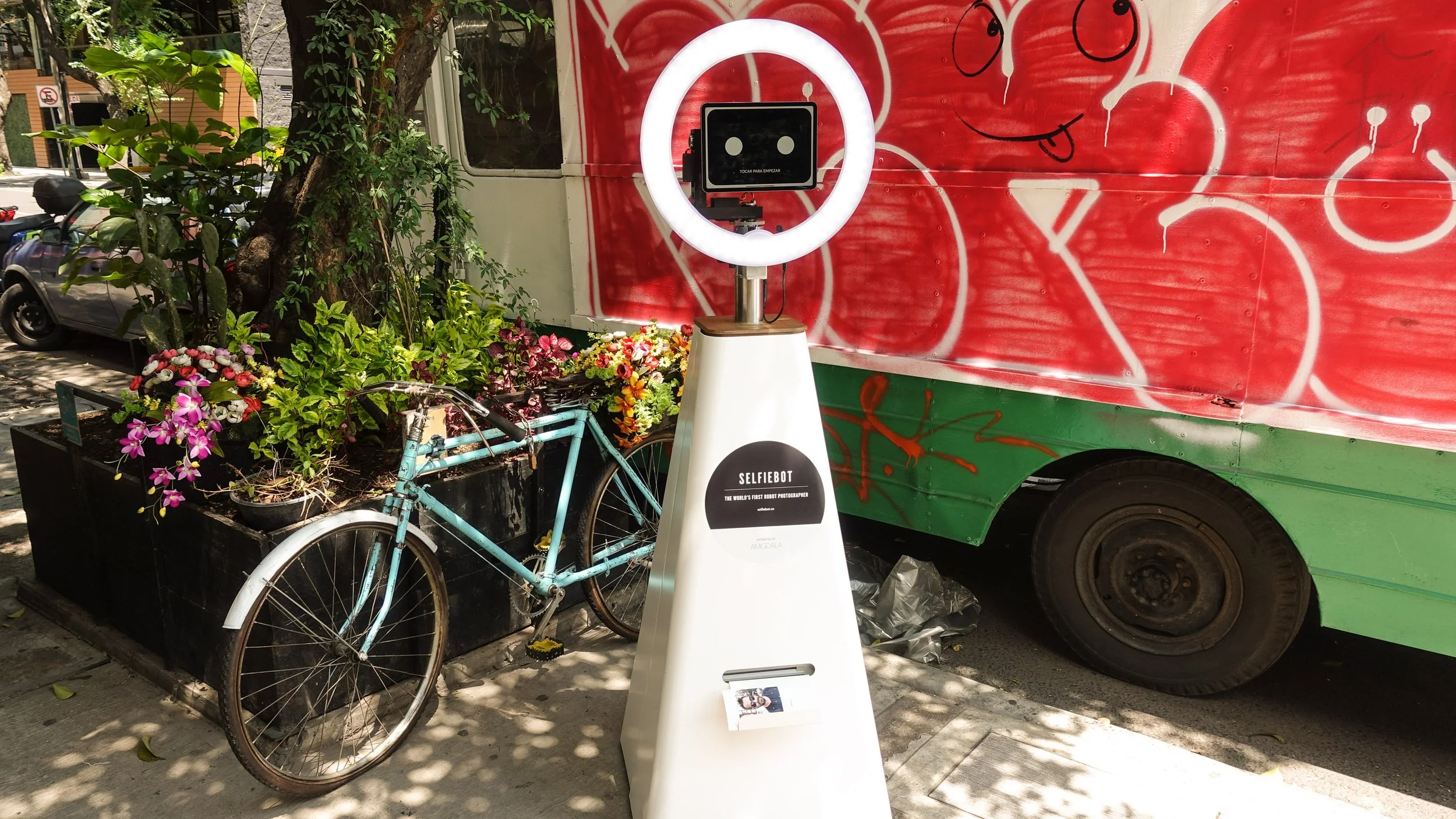 A selfie photo booth with LED ring light, a mounted tablet, and a photo strip holder is positioned on a sidewalk next to a planter with colorful flowers and a bicycle. A red graffiti wall is in the background, next to a green and red food truck.
