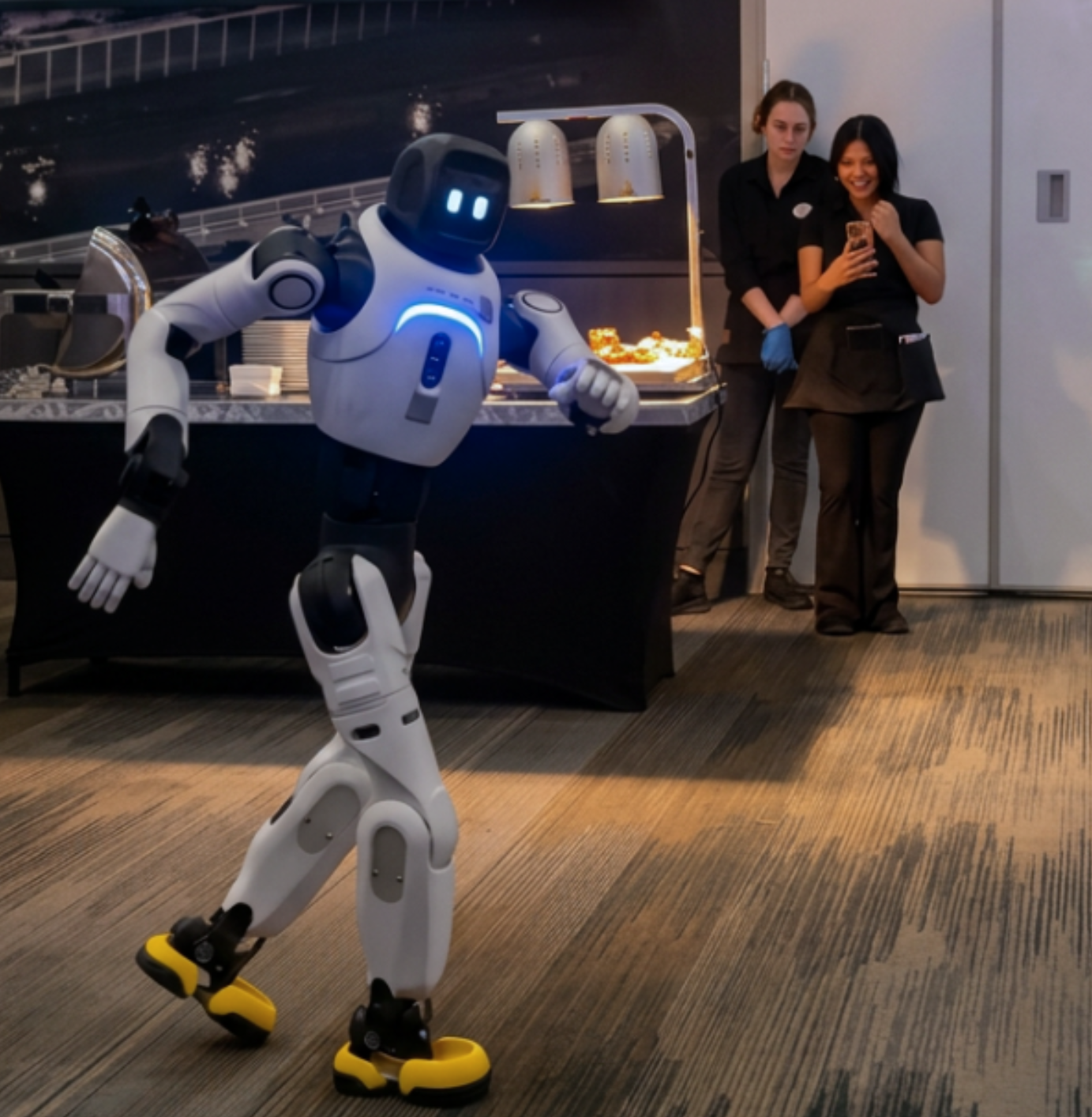Tai, a humanoid robot dancing in front of a food buffet with two women in black uniforms watching.