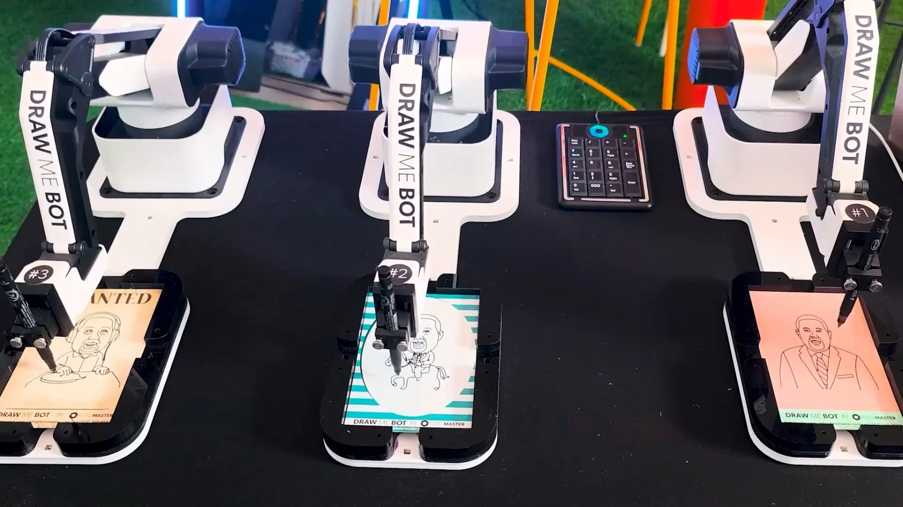 Three robotic drawing machines, each with a stylus and a screen, creating cartoon portraits of people on a table with a black cloth. The middle robot depicts a cartoon character riding a horse, while the left and right robots are drawing faces of ind