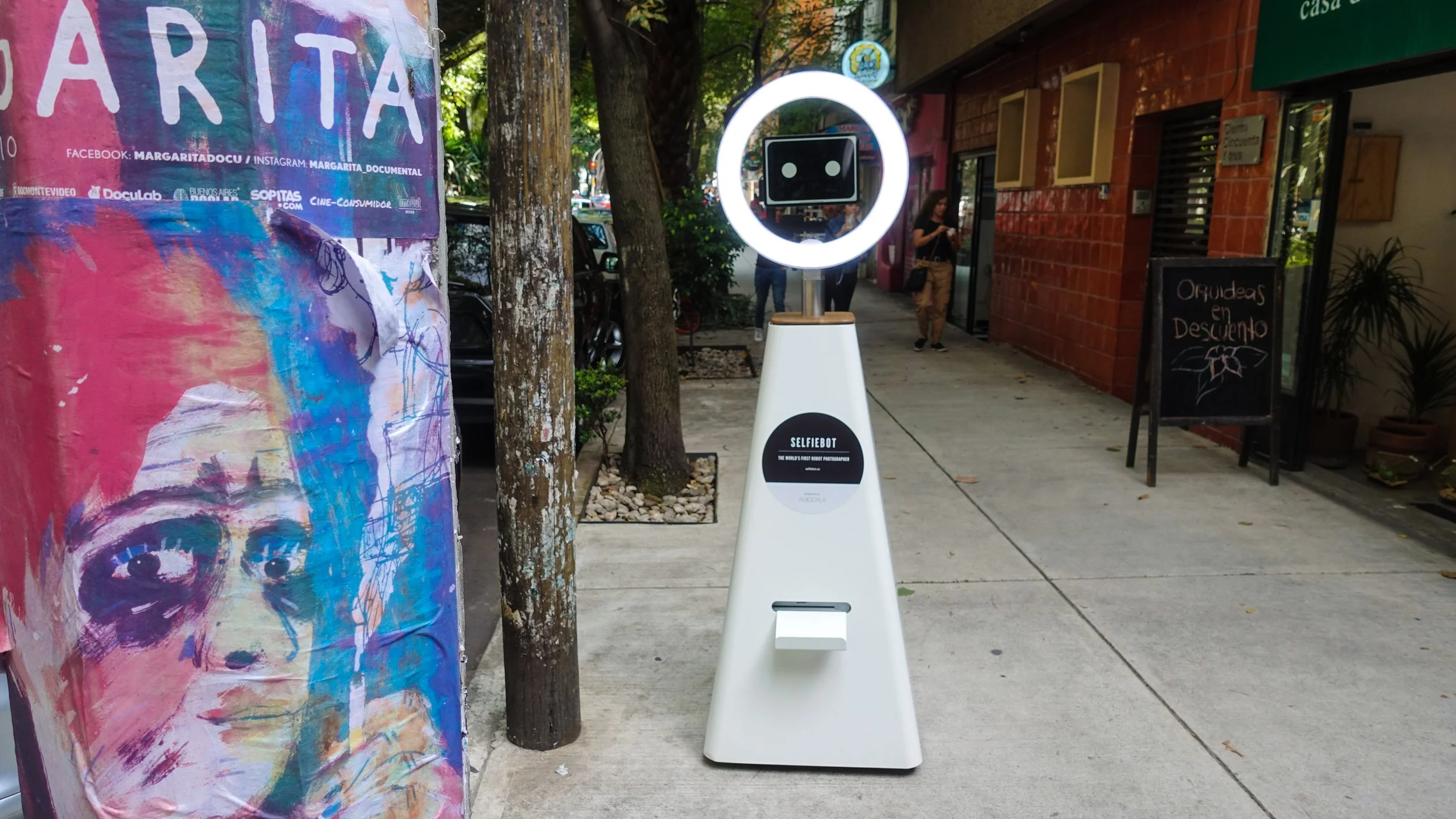 Street view featuring a selfie robot named 'Selfiebot' with a circular camera and digital face display, sidewalk artwork of a colorful face, trees, parked cars, and pedestrians.