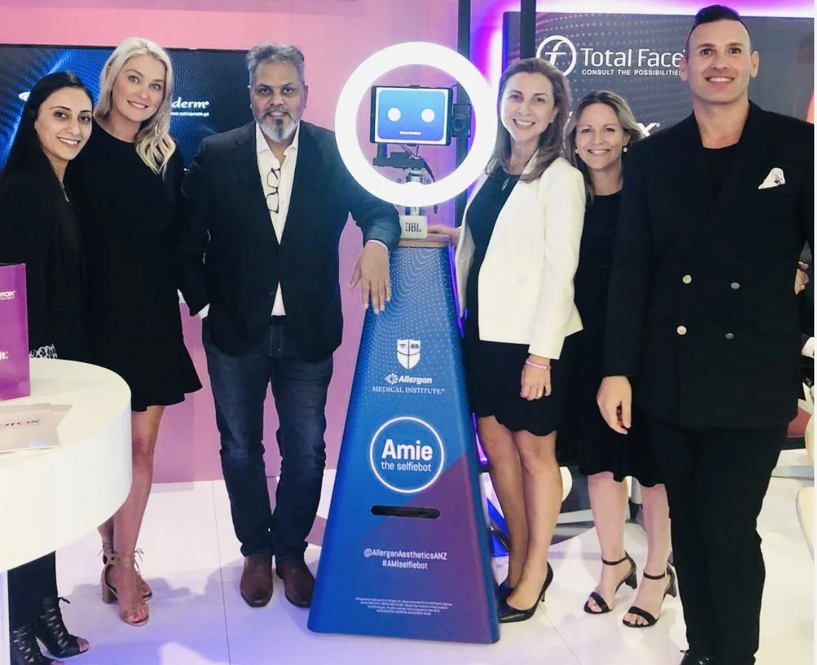 Six people standing together at an event, posing around a robot called Amie the selfiebot, with branding for Allergan Medical Institute and Total Face in the background.