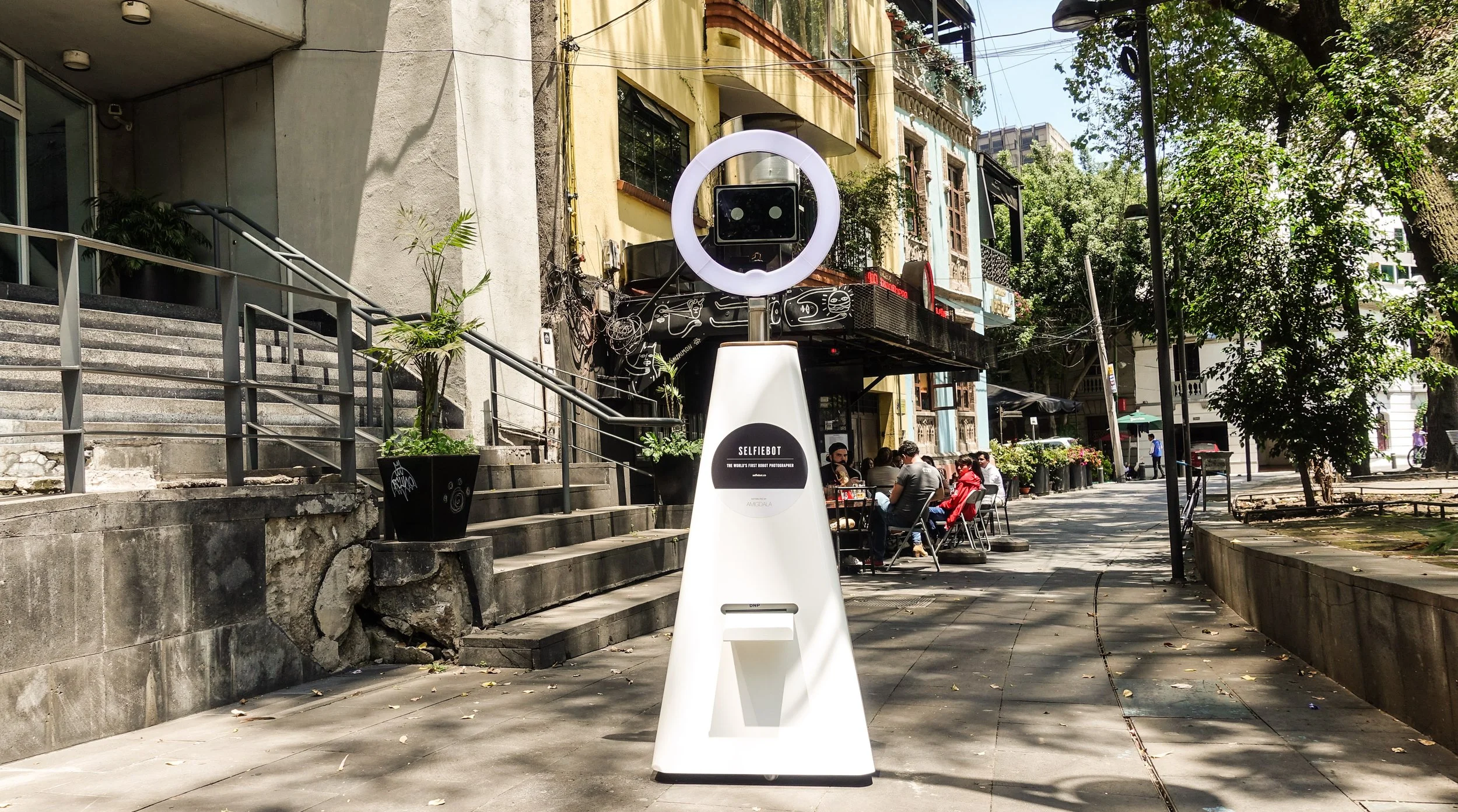 A futuristic white robot with a camera-like head and a ring light stands on a city sidewalk. In the background, people sit at outdoor tables, and buildings with storefronts and greenery are visible.