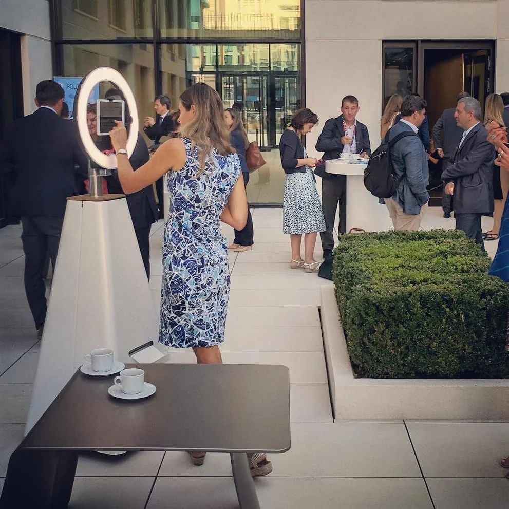 A group of people gathered outdoors at a networking event or conference, some standing and chatting, while a woman takes a photo  of herself in front of a Selfie Bot roaming robot photo booth. 