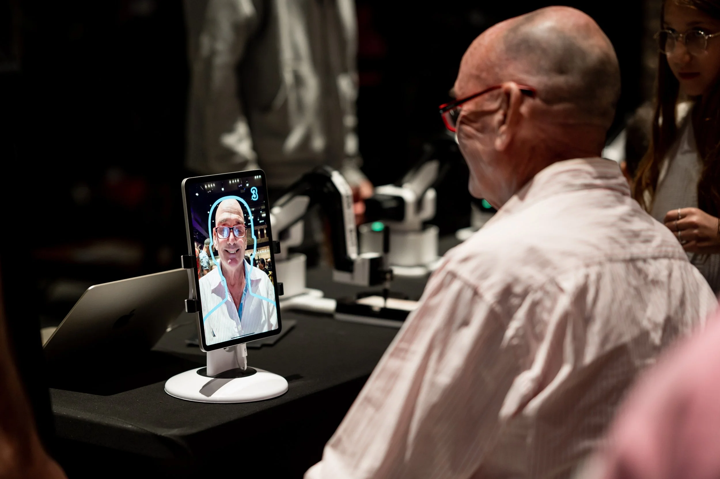 A man with glasses and a white shirt is sitting at a table, looking at a tablet displaying his own face and a digital face outline, likely during a facial recognition or security demonstration.