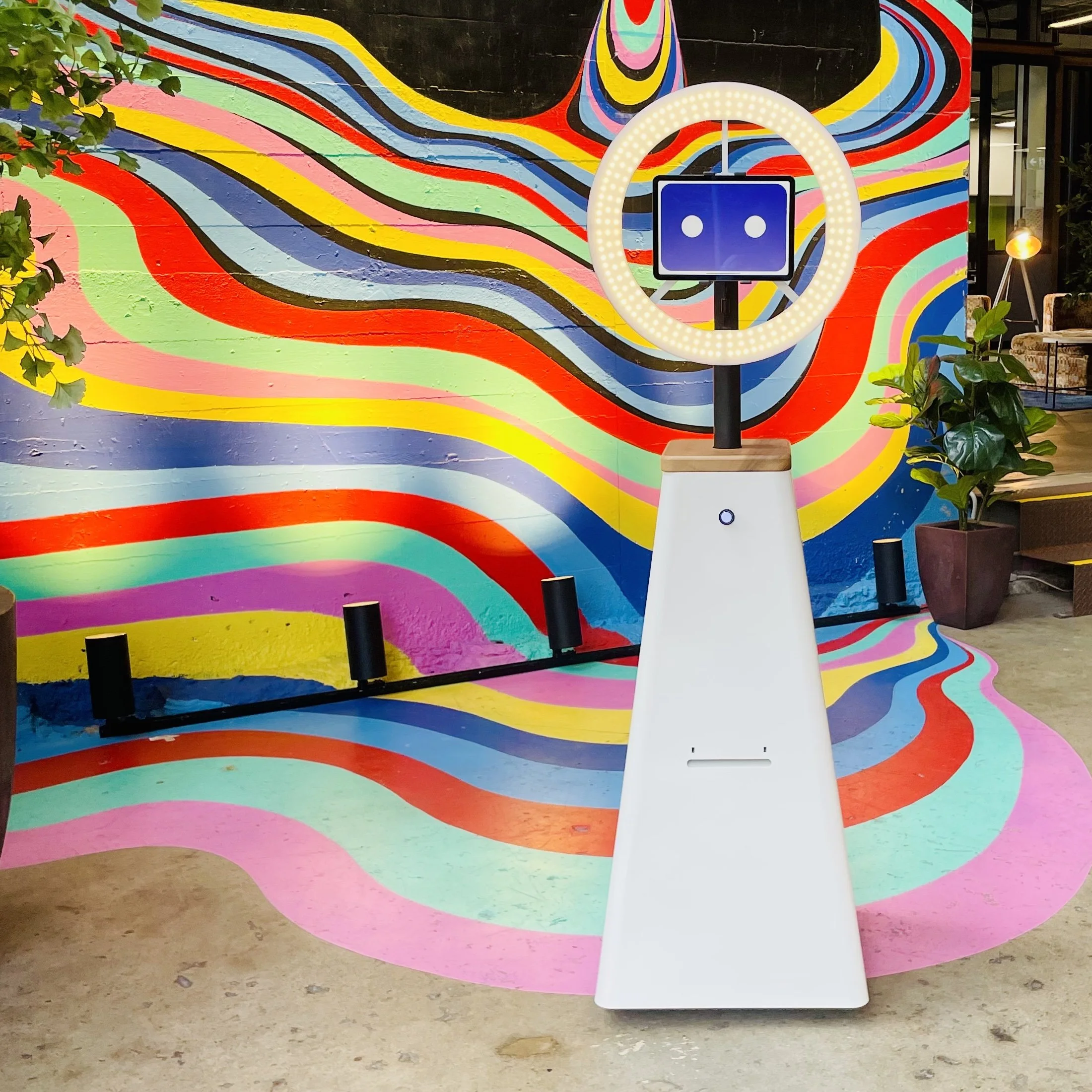A colorful indoor arcade with a vibrant, wavy, multicolored mural wall and a robot-themed basketball game with a white stand, a digital screen, and a circular ring light, surrounded by potted plants.
