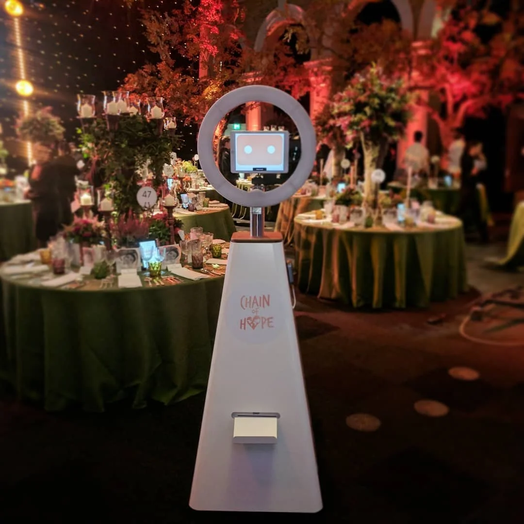 A robot with a screen face and a circular ring around its head standing in a decorated event hall with tables covered in green tablecloths, floral centerpieces, and string lights. The robot has "CHAIN OF HOPE" written on its body.