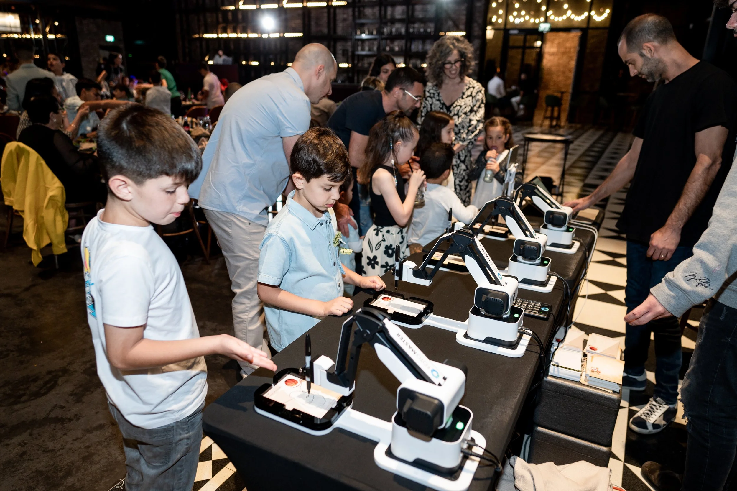 Children and adults gathered around a table with small robotic arms, some children operating the robots, in a well-lit indoor space with a checkered floor and decorative string lights.