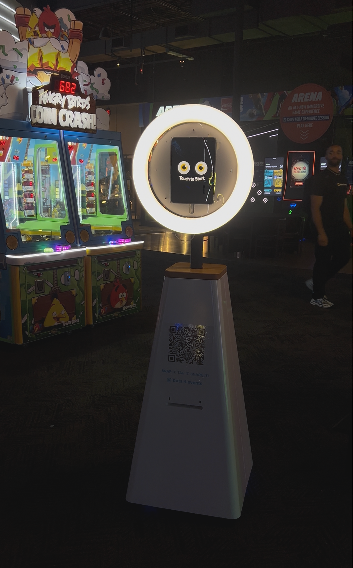 Indoor arcade with a display screen that says 'Touch to Start' surrounded by a circular light. To the left, there are two Angry Birds-themed claw machines. A sign at the top reads 'Angry Birds Coin Crash!' with a bonus score display. A person is standing in the background near other arcade machines and signs.