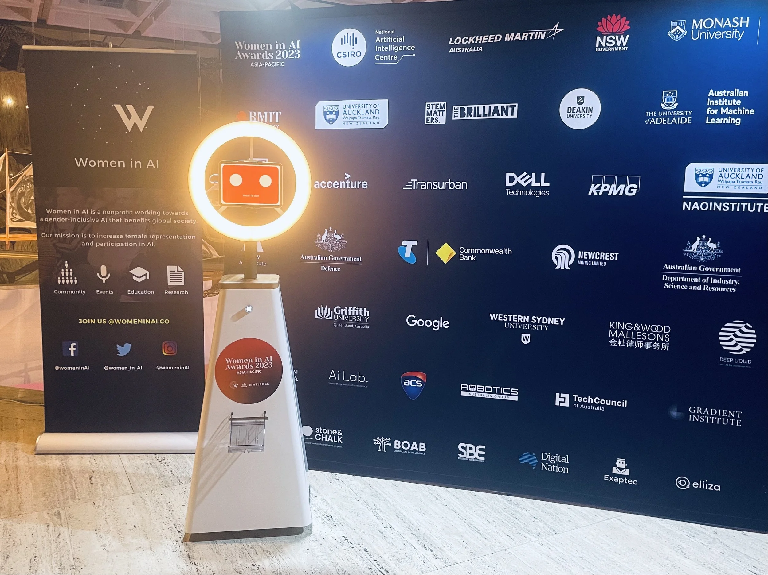 Large blue backdrop with logos of various sponsors and organizations for the Women in AI Awards 2023 Asia-Pacific, including university and tech company logos. To the left, a black vertical banner with the Women in AI logo, a brief description, and s