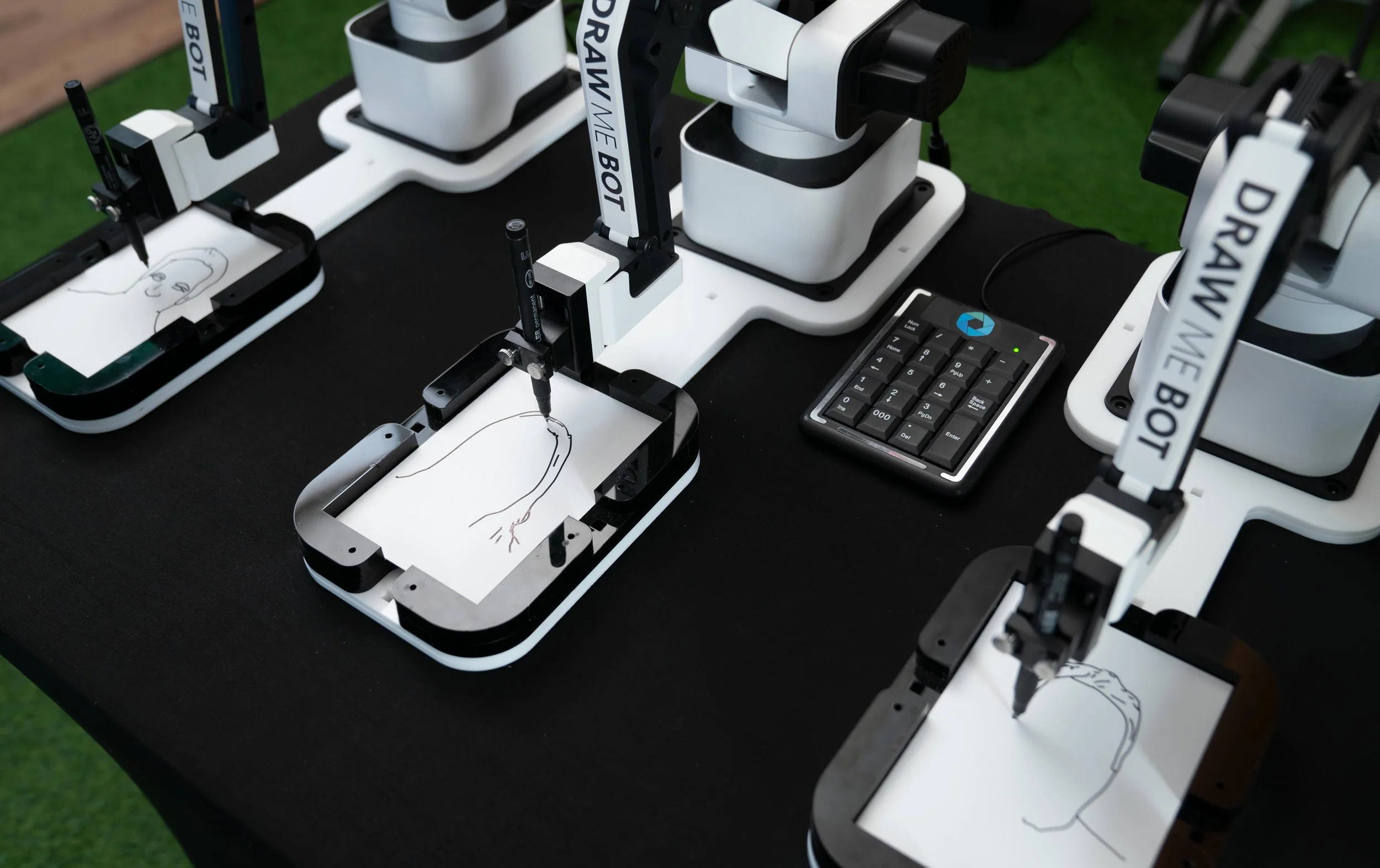 Multiple robotic arms drawing digital portraits on tablets, with a calculator on a black table.