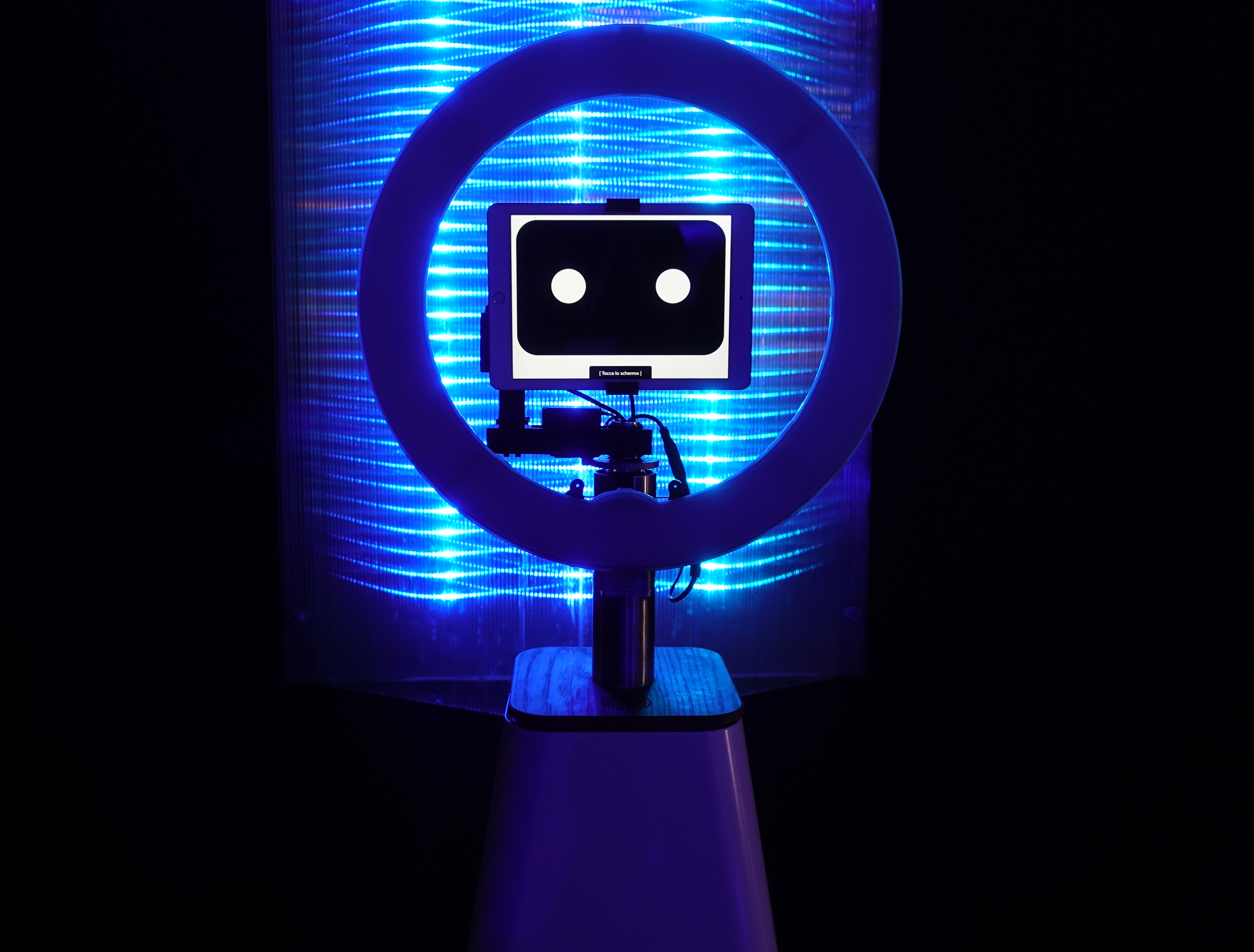 A robot with a screen displaying two dots inside a ring, illuminated by blue LED lights against a black background.