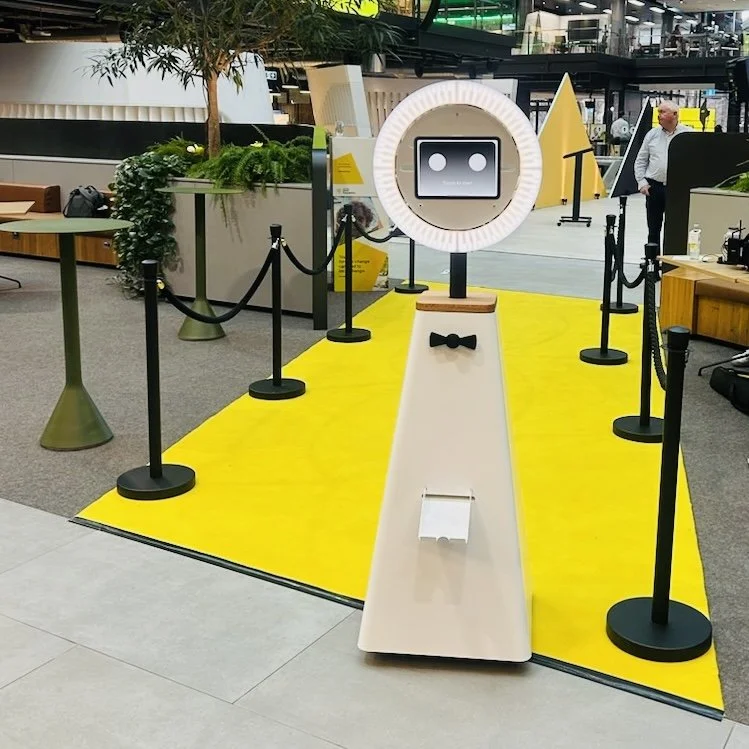 A photobooth setup with a robot face on a screen, surrounded by a ring light, on a white ramp with a yellow carpet, velvet ropes, in an indoor public space, possibly an airport or mall.