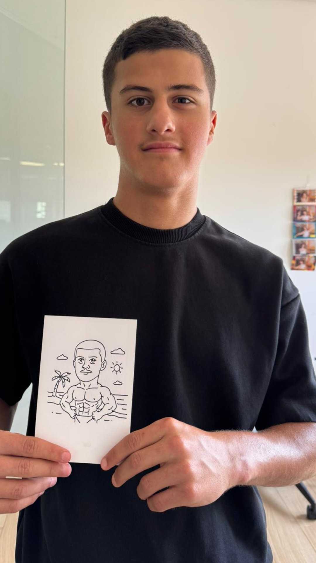 A young man with short brown hair and fair skin is standing indoors, holding a drawing of a muscular man with a short haircut on a piece of paper. The drawing is set against a background with a palm tree, clouds, and the sun.