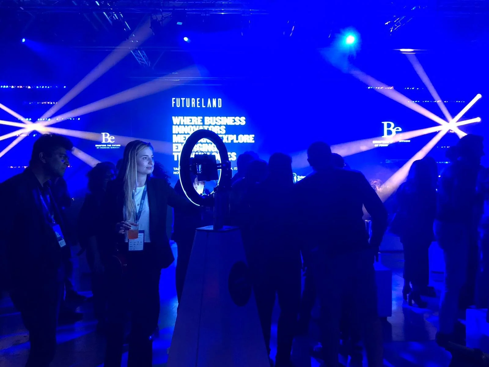 People attending a conference or event, standing in front of a blue-lit stage with large screens displaying the words 'FUTURELAND' and motivational phrases, with bright stage lights and a circular device in the foreground.