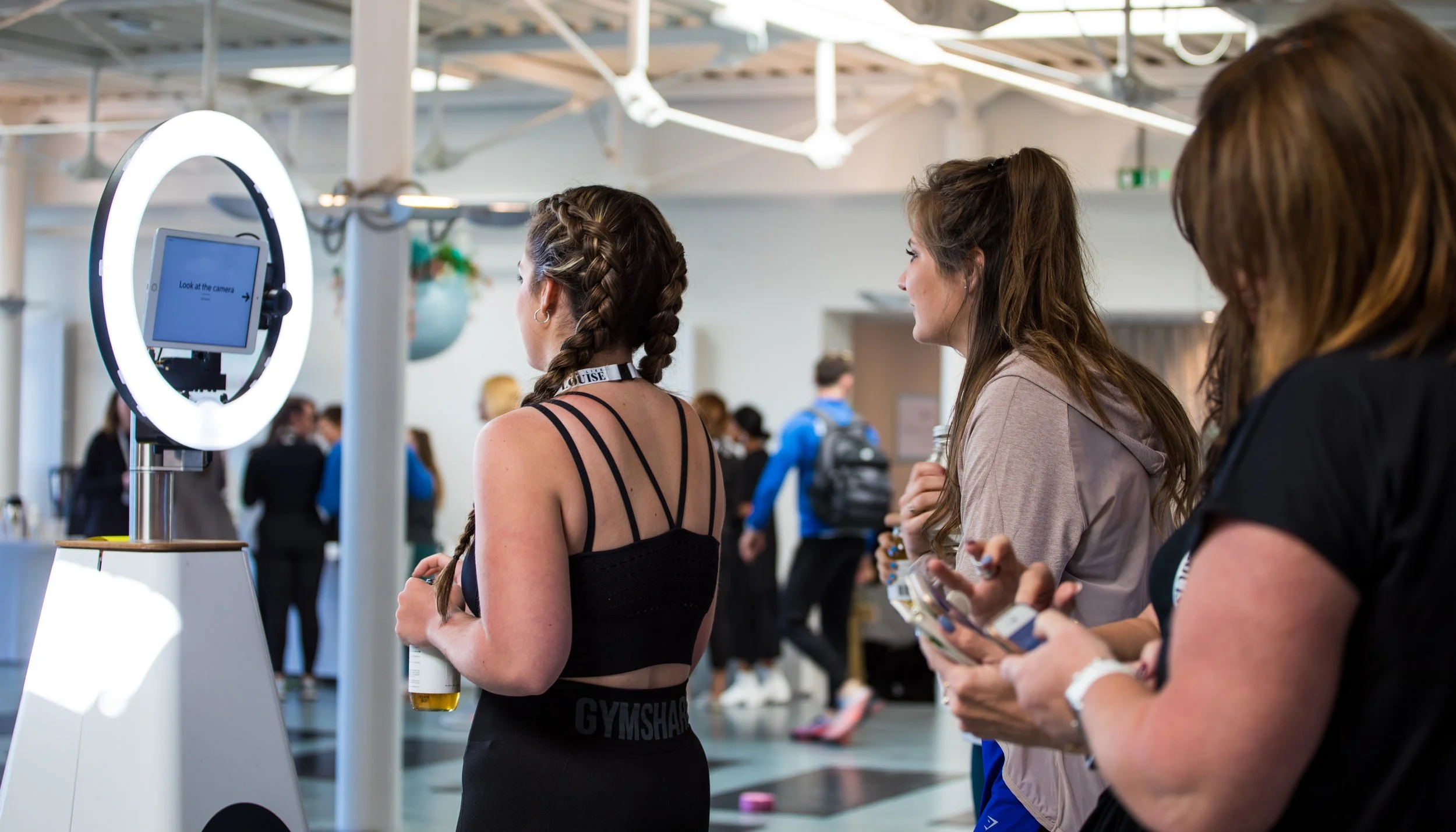 People standing in line at a registration or check-in area in an indoor event space, using a digital kiosk with a ring light, with others in the background.