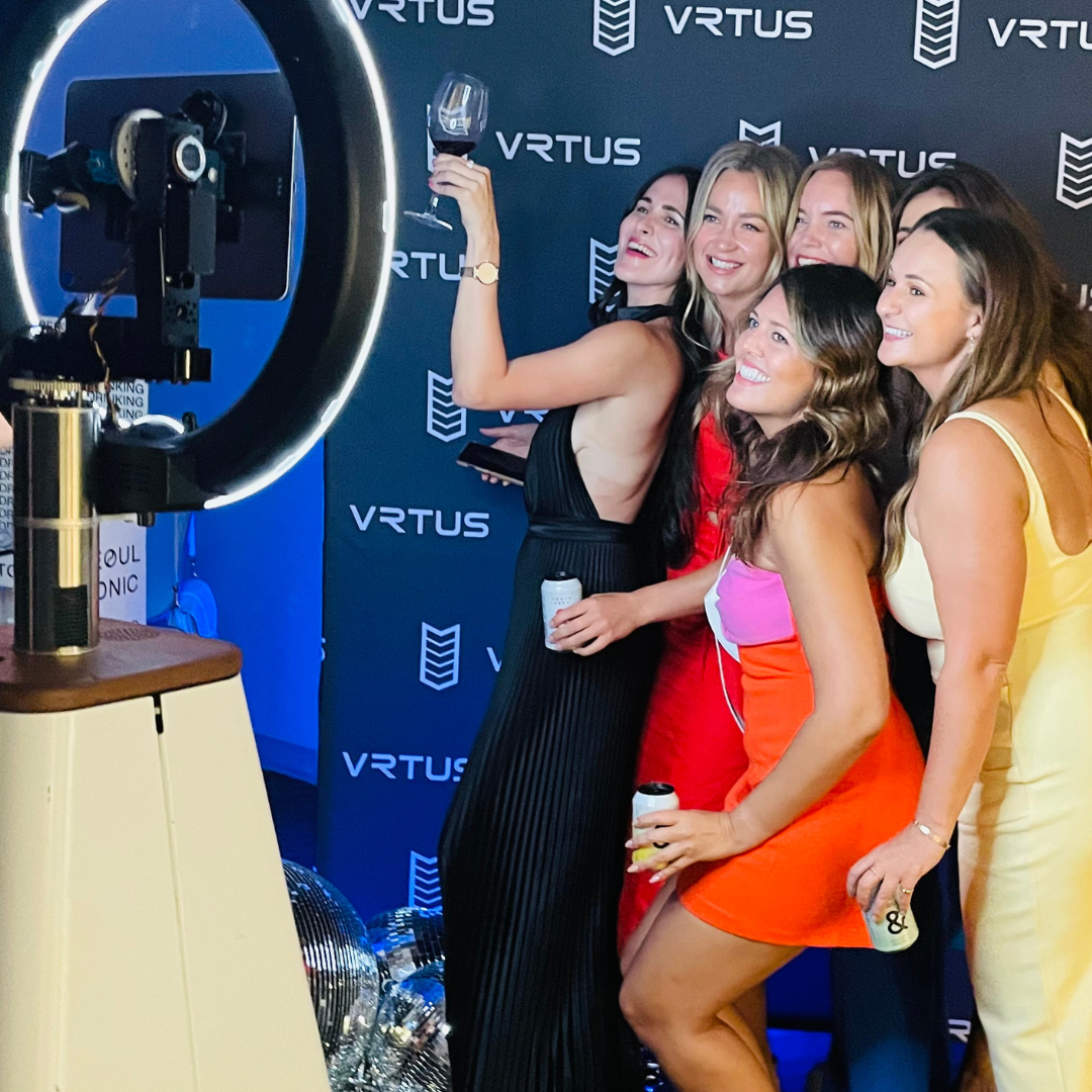 A group of women taking a selfie at a party, with a person holding a wine glass in the background.