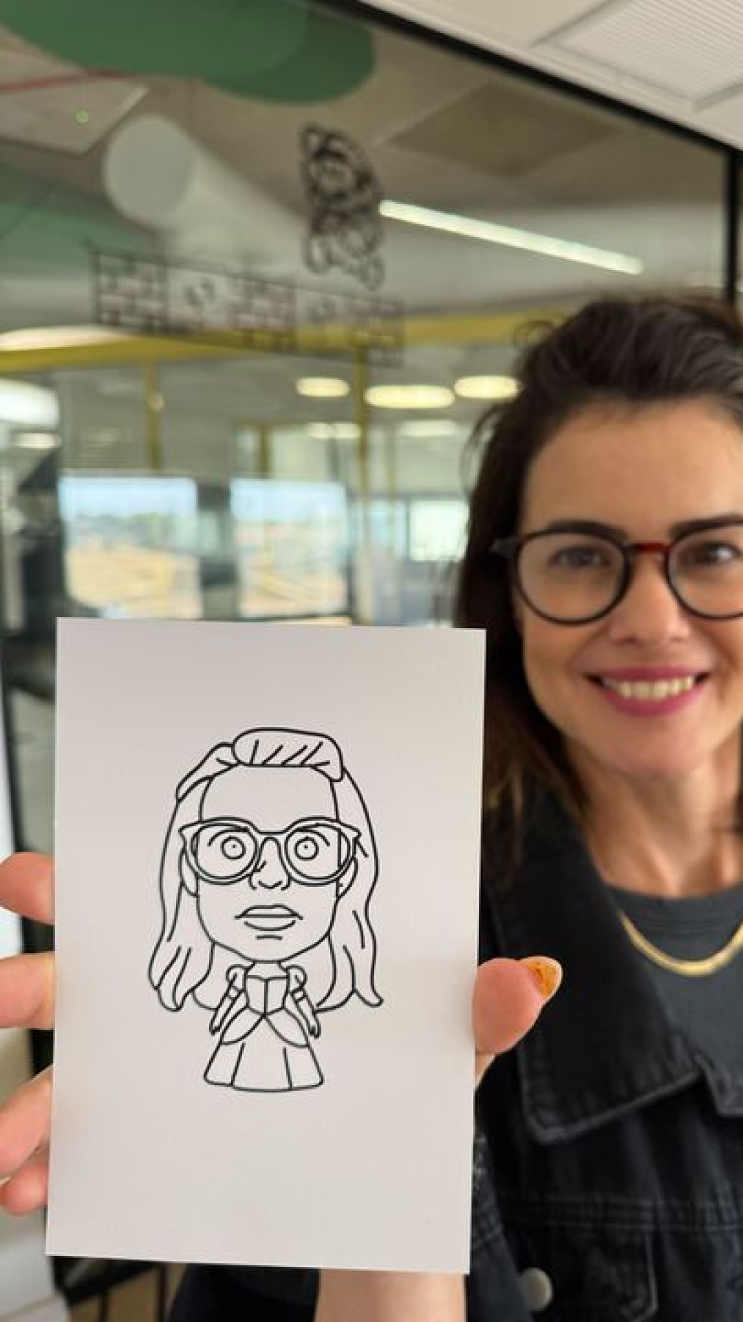 A woman with glasses and a smile holding a drawing of herself in front of her, with a woman with glasses and curly hair in the background.