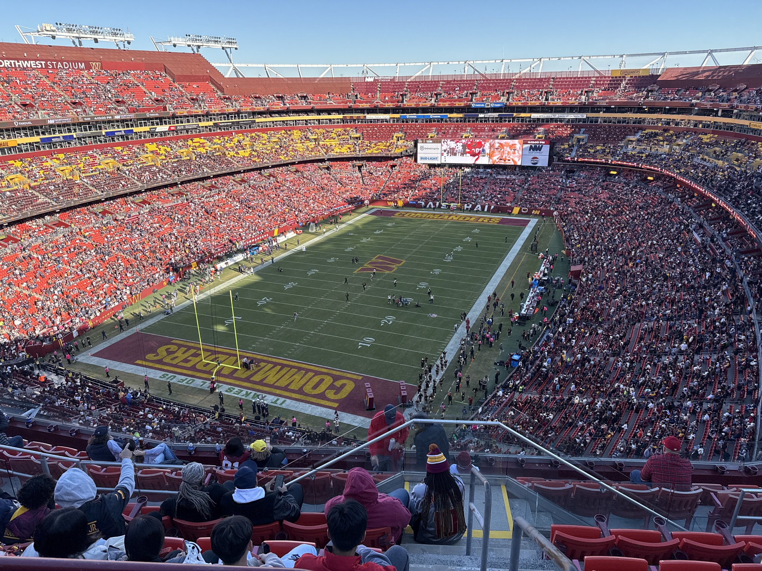 Wide view of a football stadium filled with spectators during a game, with players on the field and large screens displaying game footage.