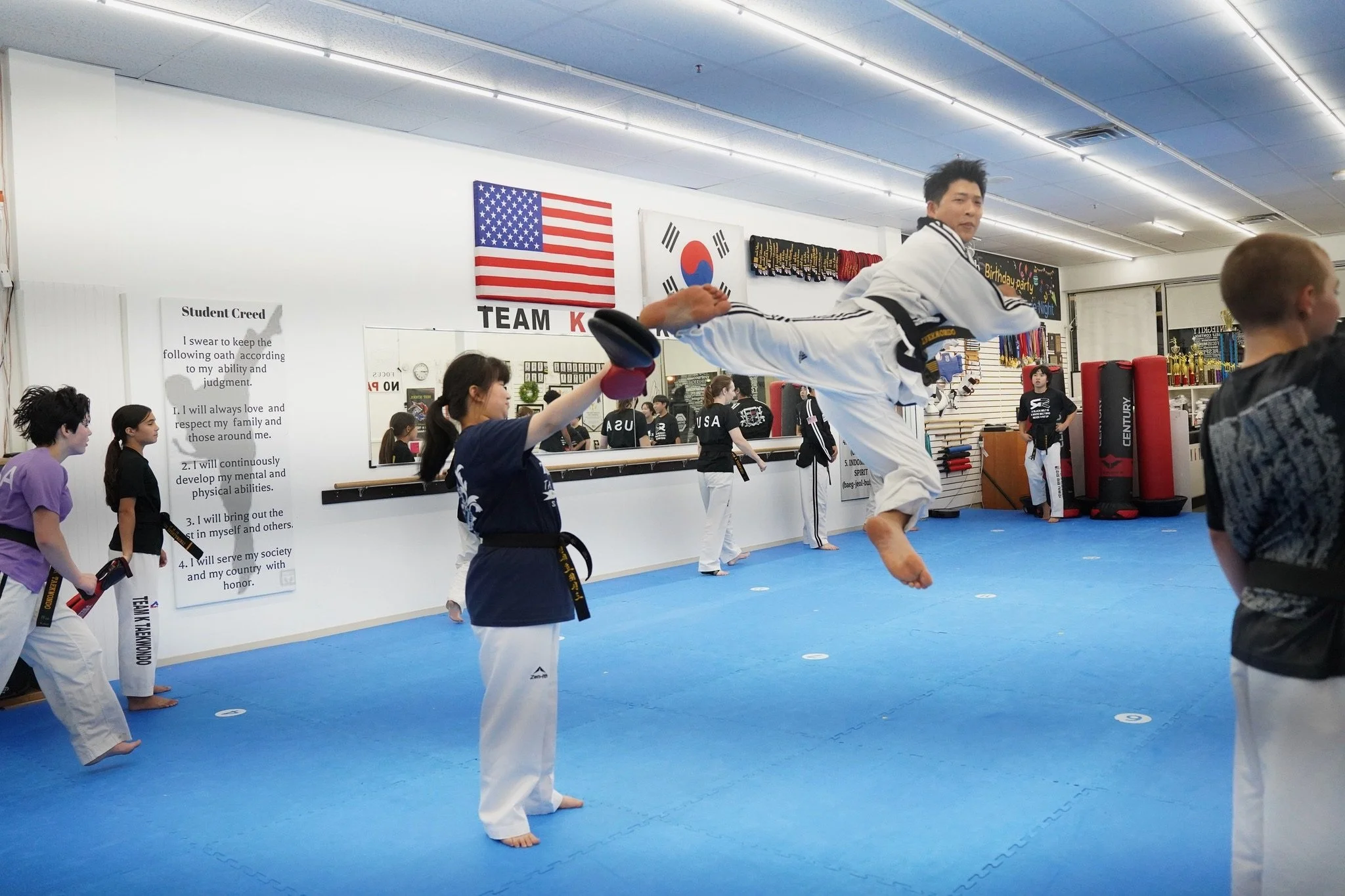 Martial arts class practicing kicks and punches in a martial arts dojo with American and Korean flags on the wall.