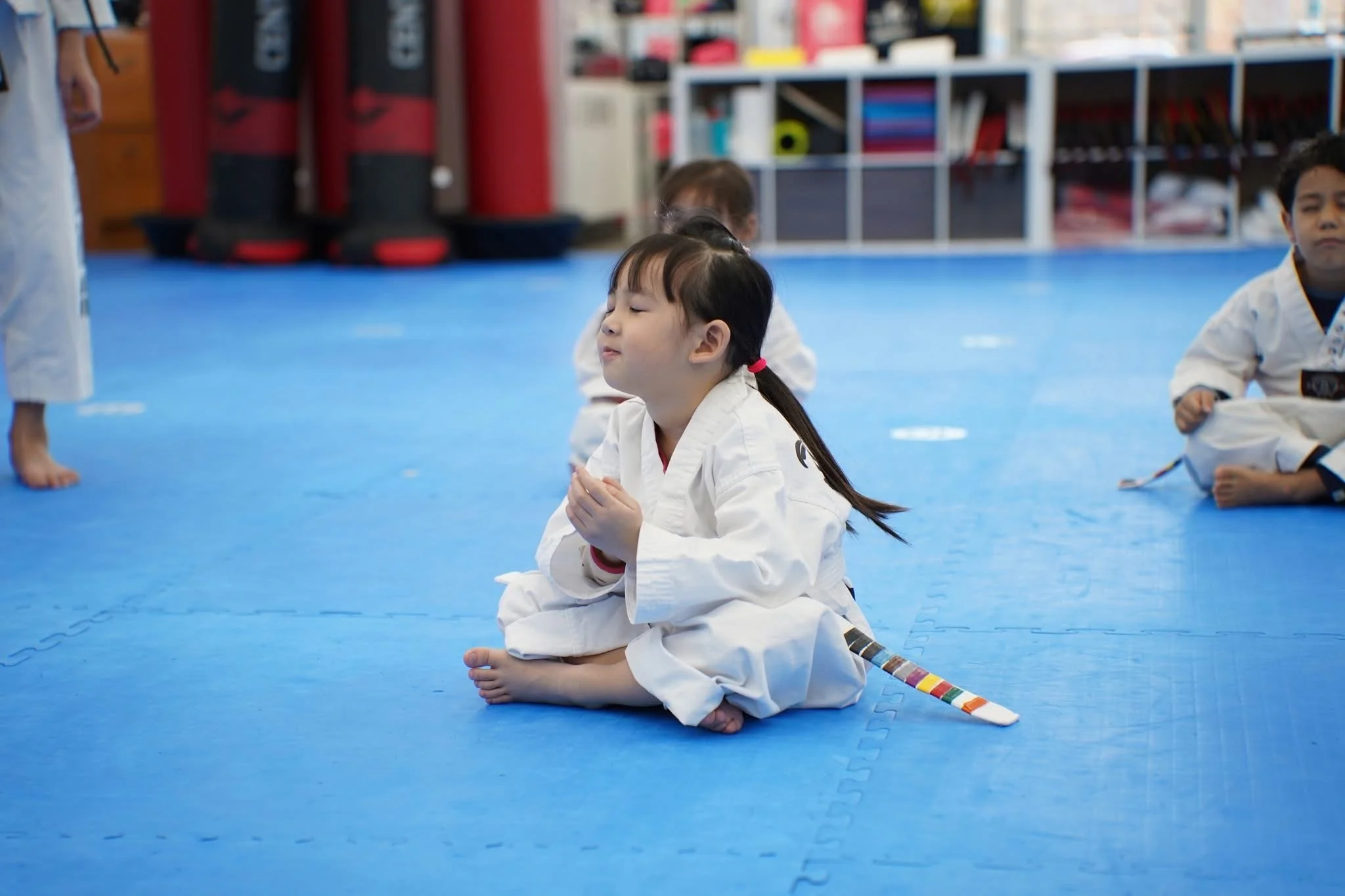A young girl in a white martial arts uniform sitting cross-legged on a blue mat, with her eyes closed and hands in a prayer position, while other children in similar uniforms practice in the background.