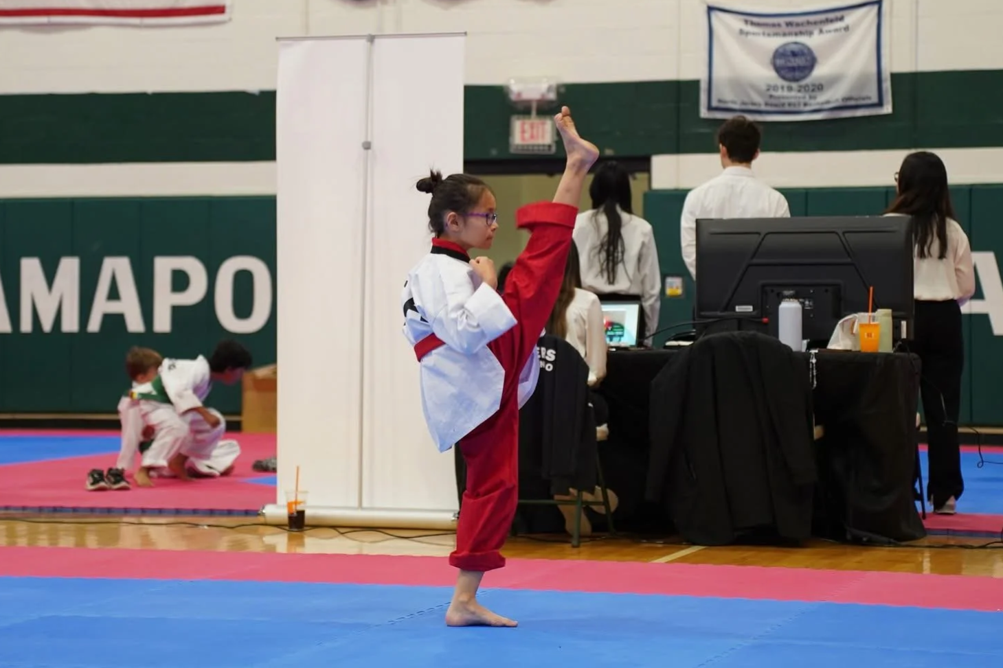A young girl in a taekwondo gi practicing a high kick at a martial arts competition in a gymnasium, with other competitors and officials in the background.