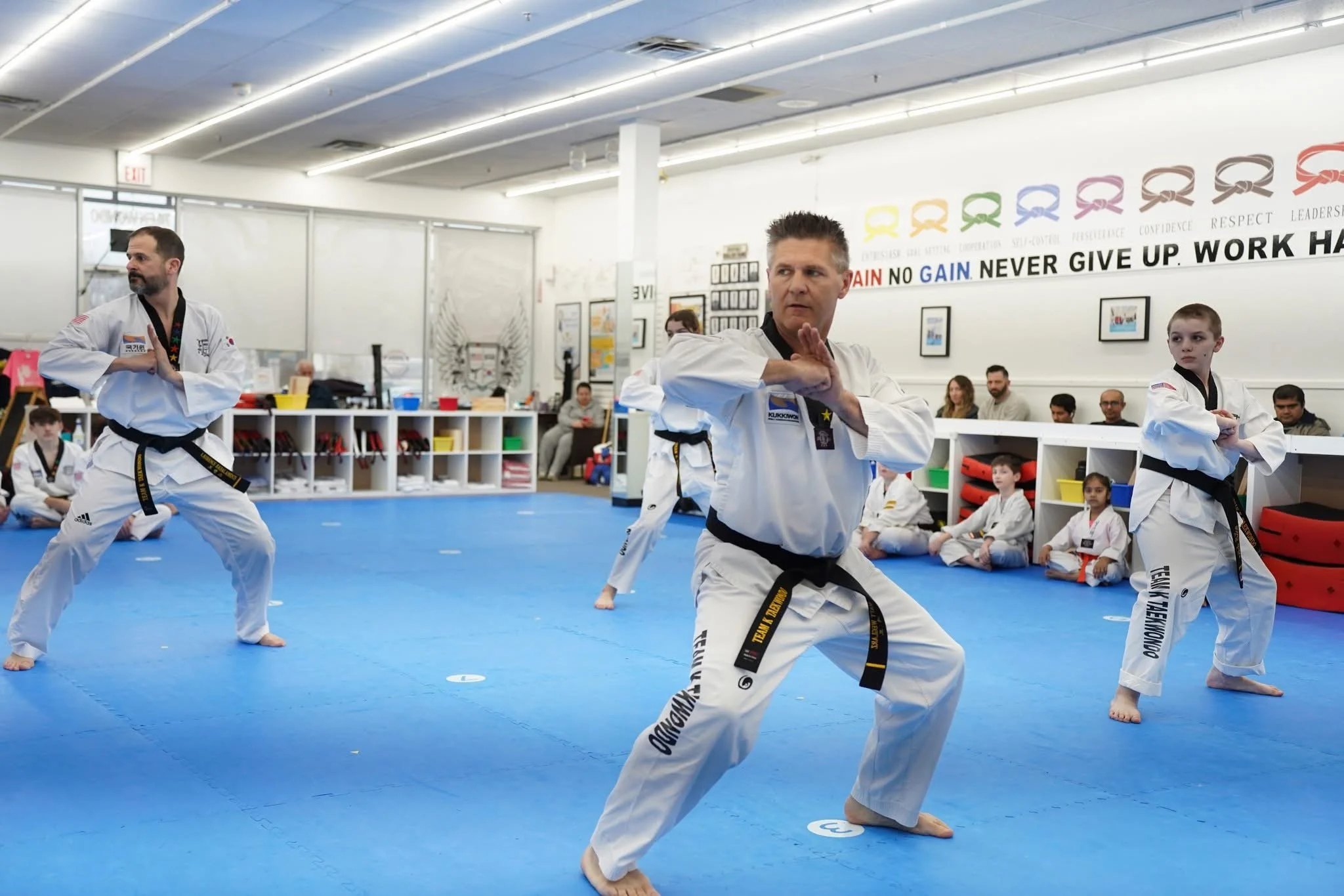 Martial arts class with young and adult students in white uniforms practicing moves on blue mats, with colorful motivational banners and seated students observing in the background.