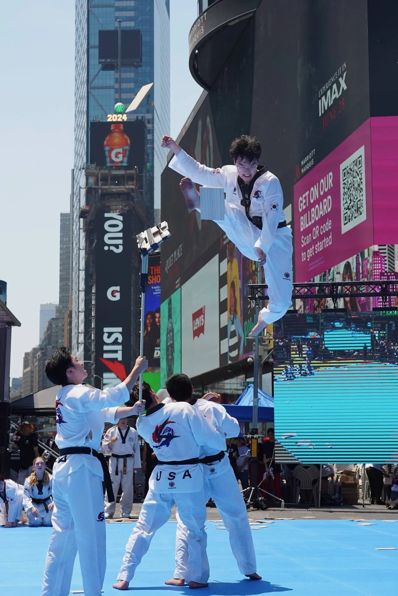 Martial artists practicing Taekwondo in an outdoor urban setting with bright blue mat. Participants wear white uniforms with an American flag on the sleeve and
